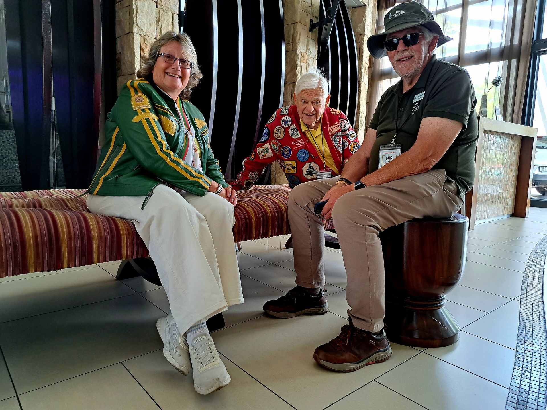 Three people seated on a bench indoors, wearing colorful clothing, with a tiled floor and striped backdrop.