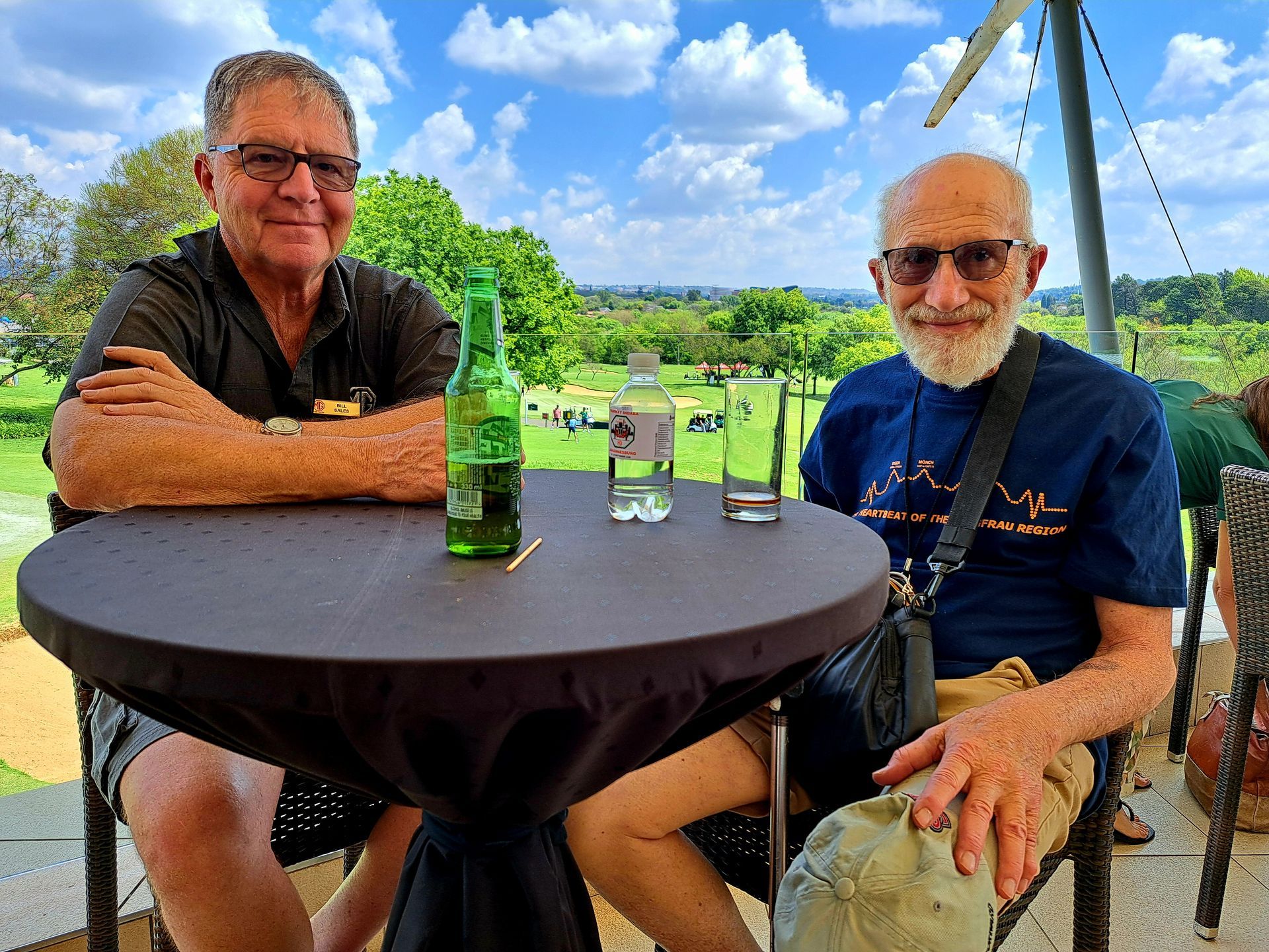 Two men seated at an outdoor table with drinks, smiling under a patio with a green lawn behind them