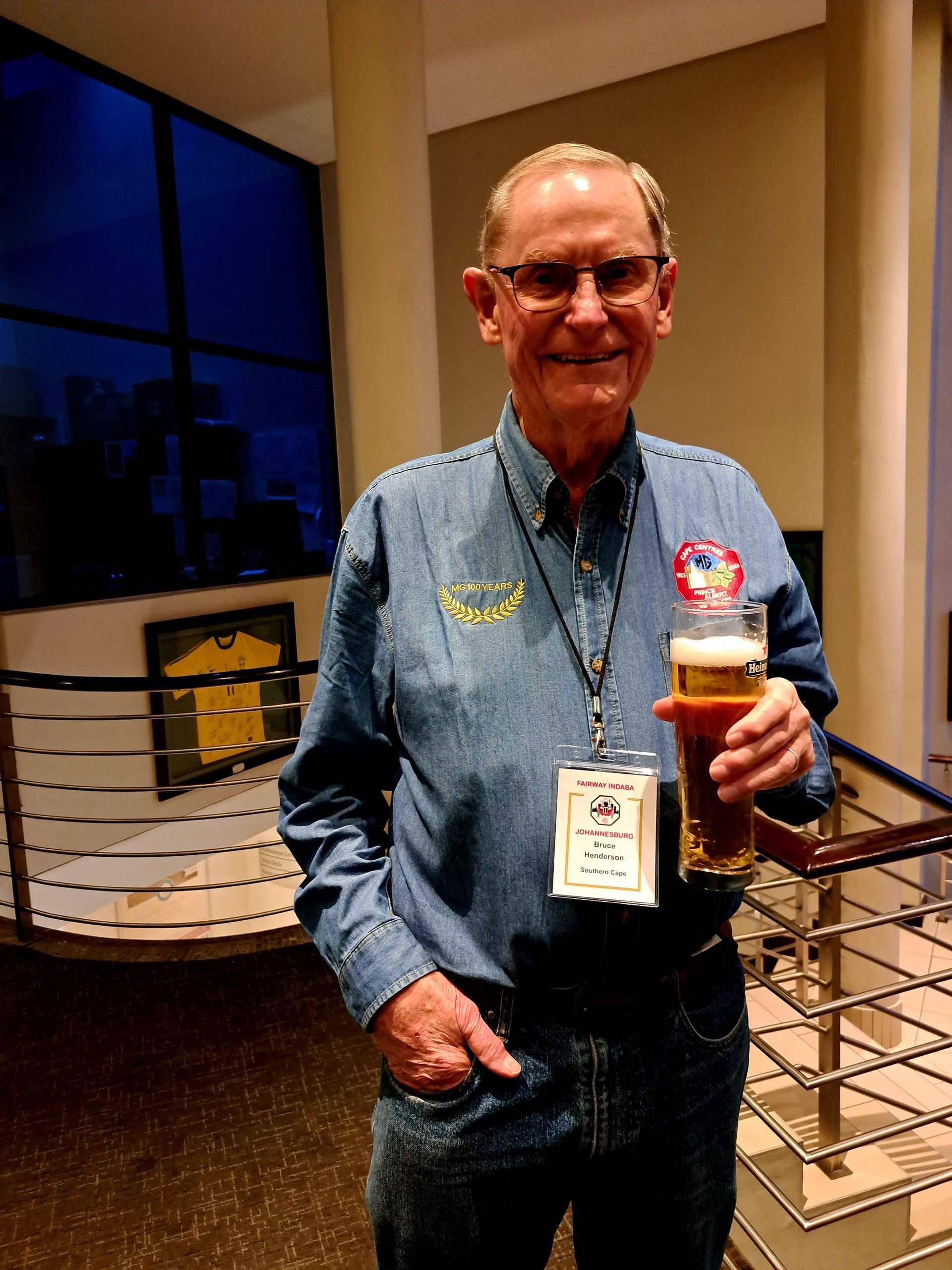 Man standing indoors holding a beer, wearing a denim shirt and name badge, near windows and railings.