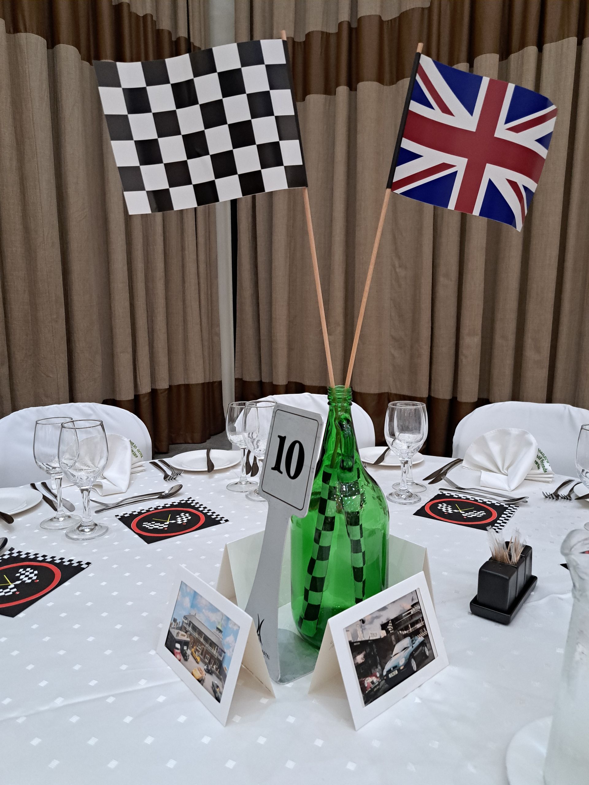 Race-themed table centerpiece with checkered and British flags, green bottle vase, and framed photos on a white tablecloth