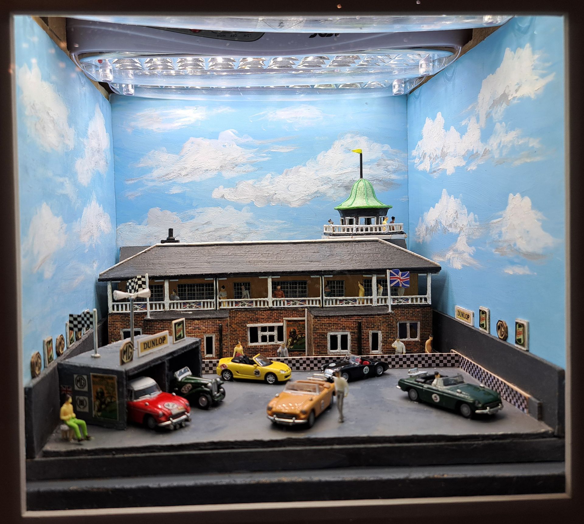 Miniature model of a brick building and parking lot with toy cars under a cloudy blue sky backdrop