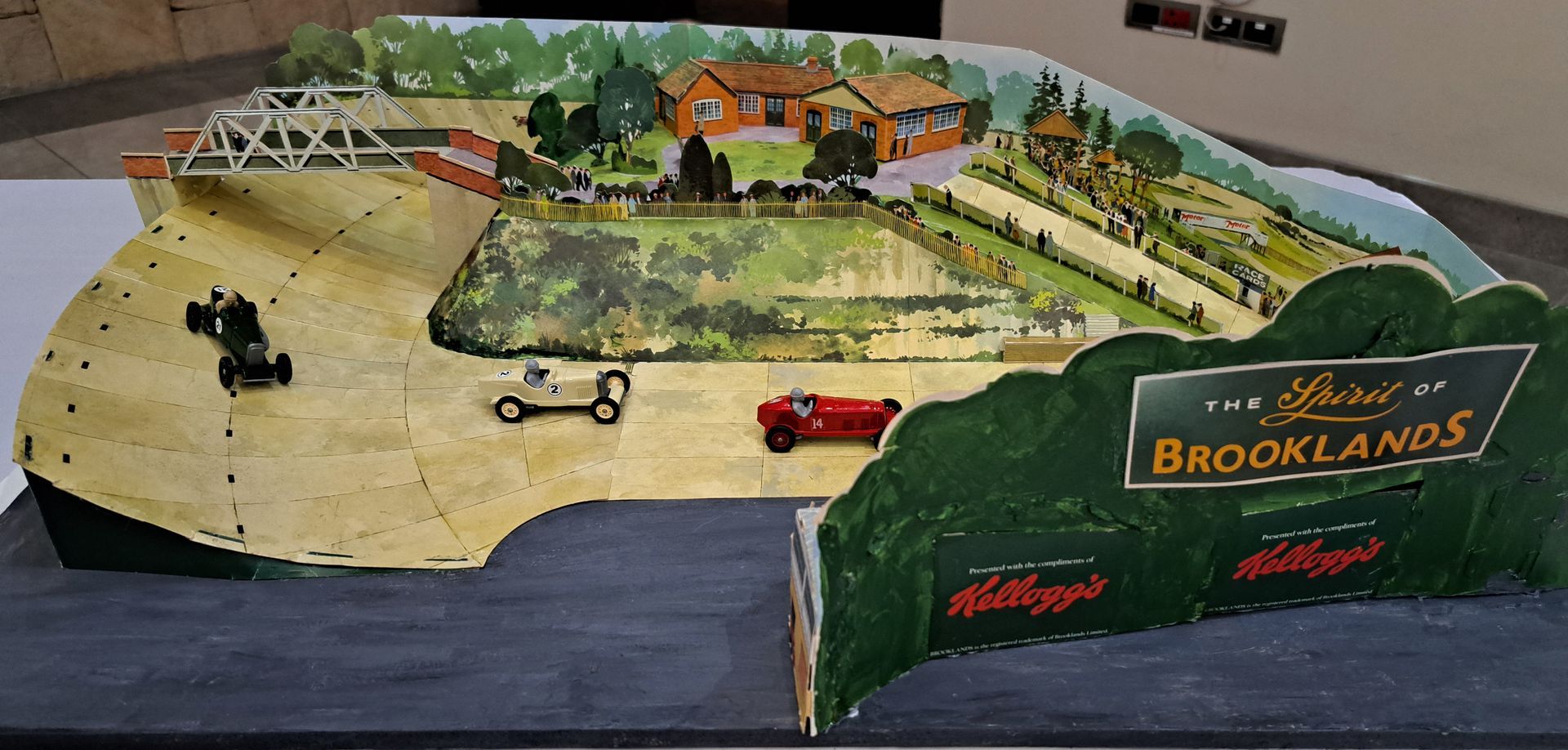 Scale model of Brooklands racetrack with green infield, small buildings, cars, and a hillside roadway