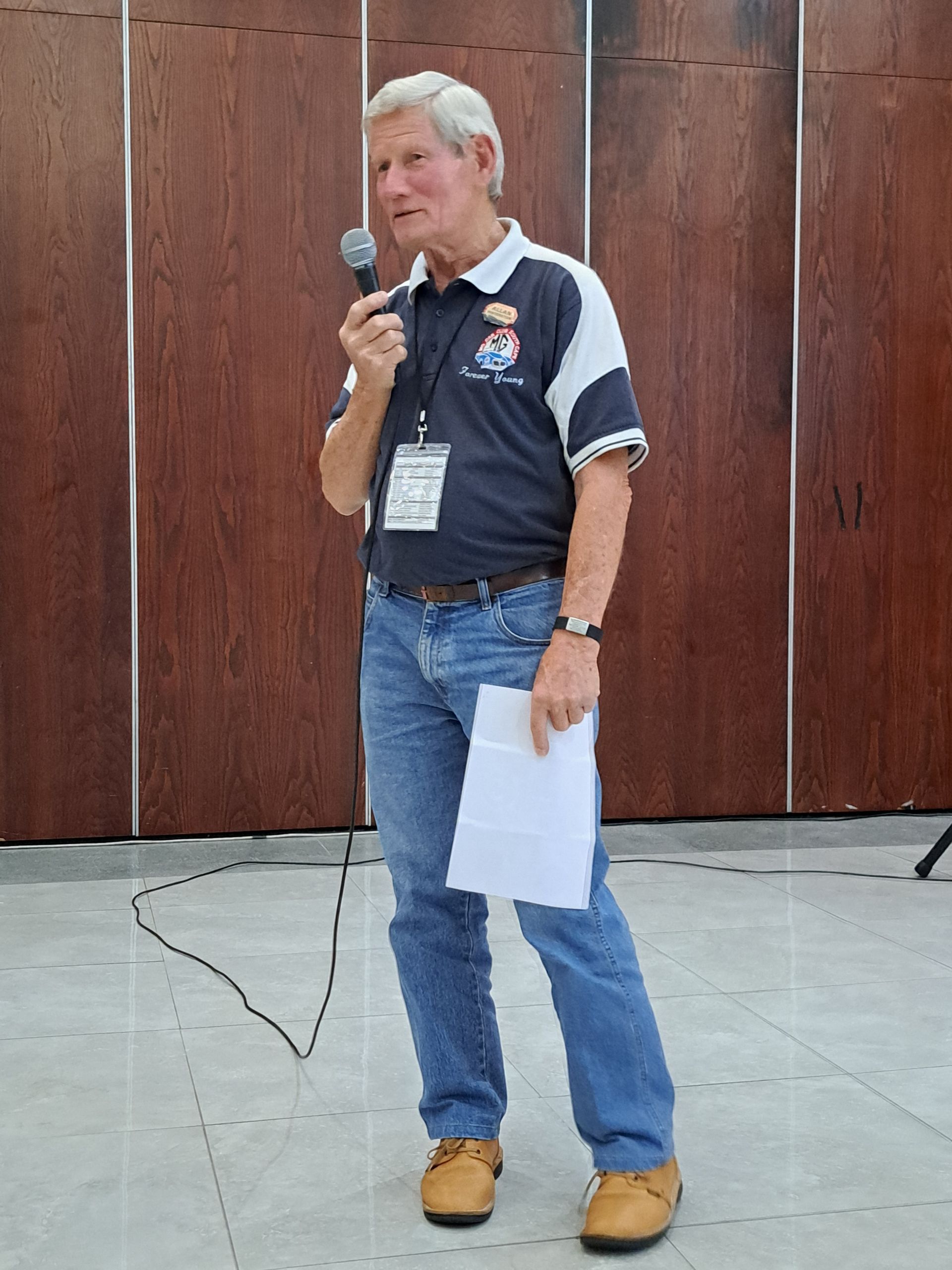 Man speaking into a microphone, holding papers, standing indoors in front of wood-paneled walls.