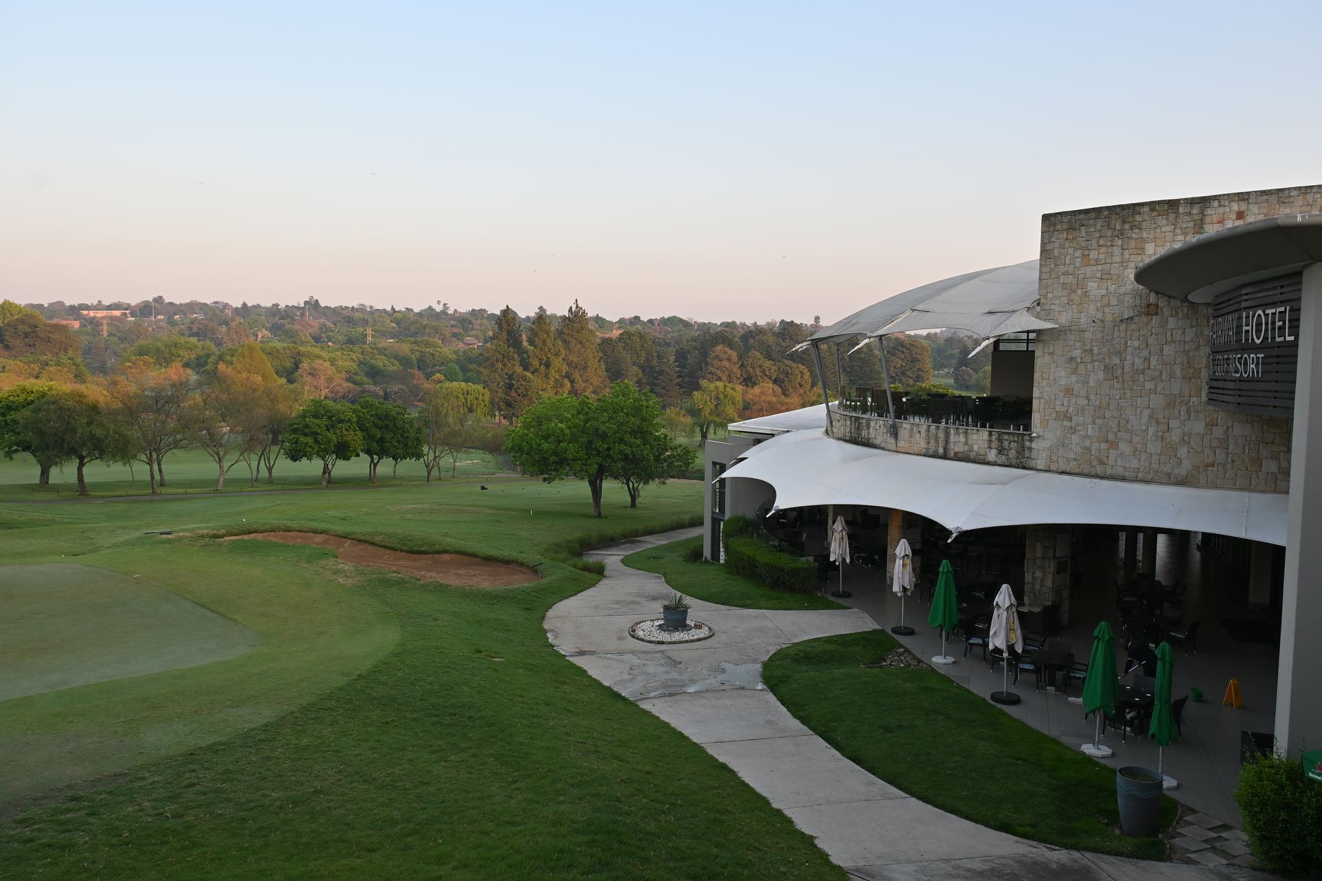 Golf course beside a curved resort building at sunset, with trees and a patio walkway.