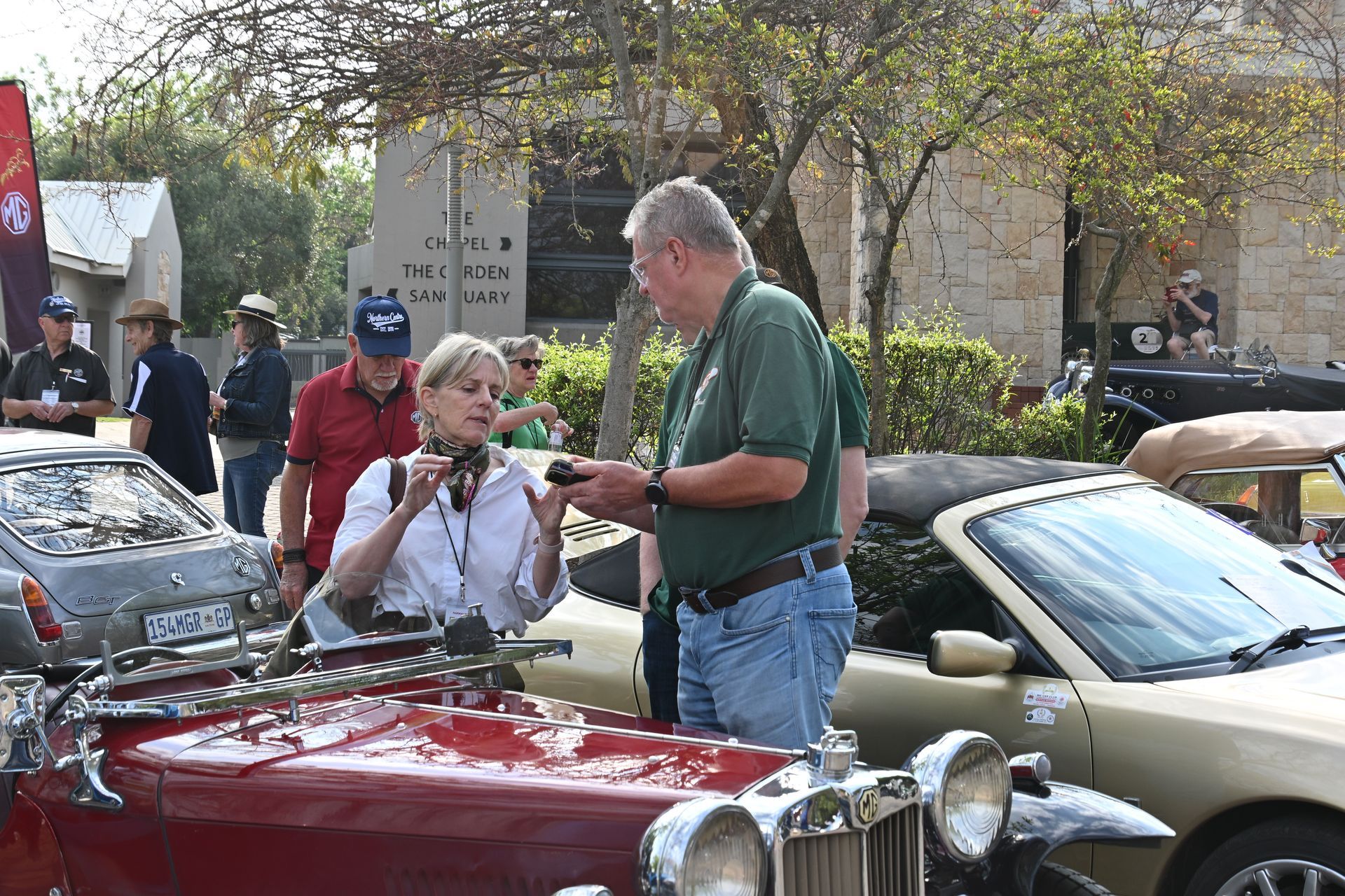 People chatting beside a red vintage car at an outdoor car show, with other classic cars nearby.