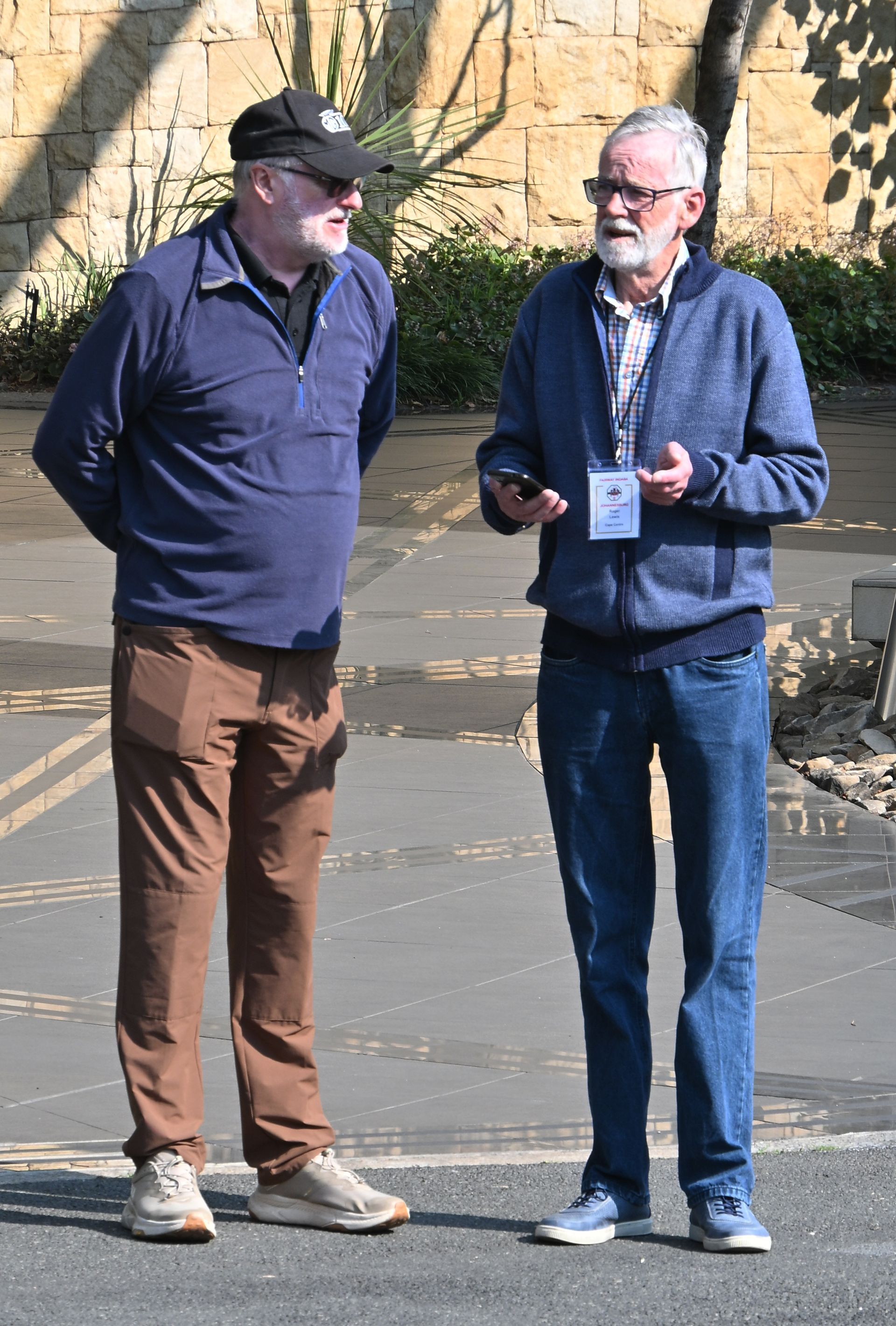 Two people stand talking outdoors on a paved walkway, one holding a phone and wearing a lanyard.