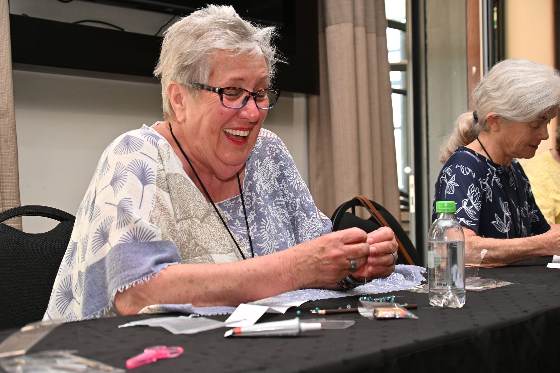Two women seated at a table, smiling and working with small items in a meeting room.