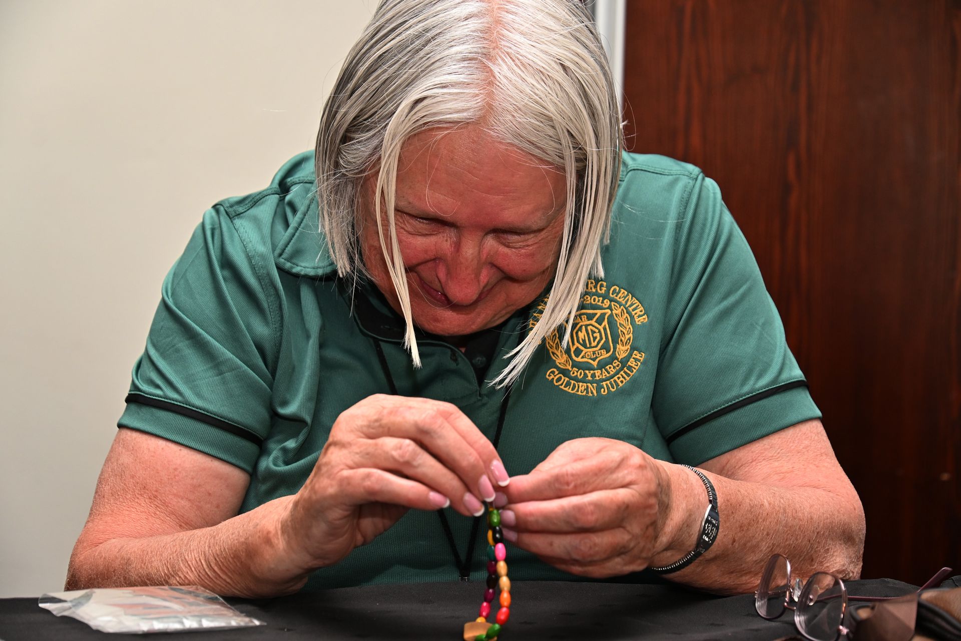 Older person in a green shirt threading colorful beads at a table