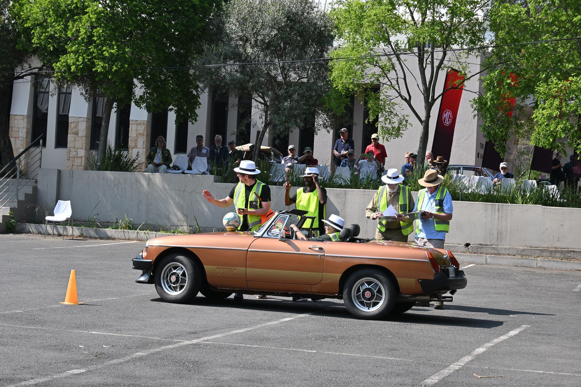 Orange convertible with several flaggers in neon vests standing behind it on a street course.