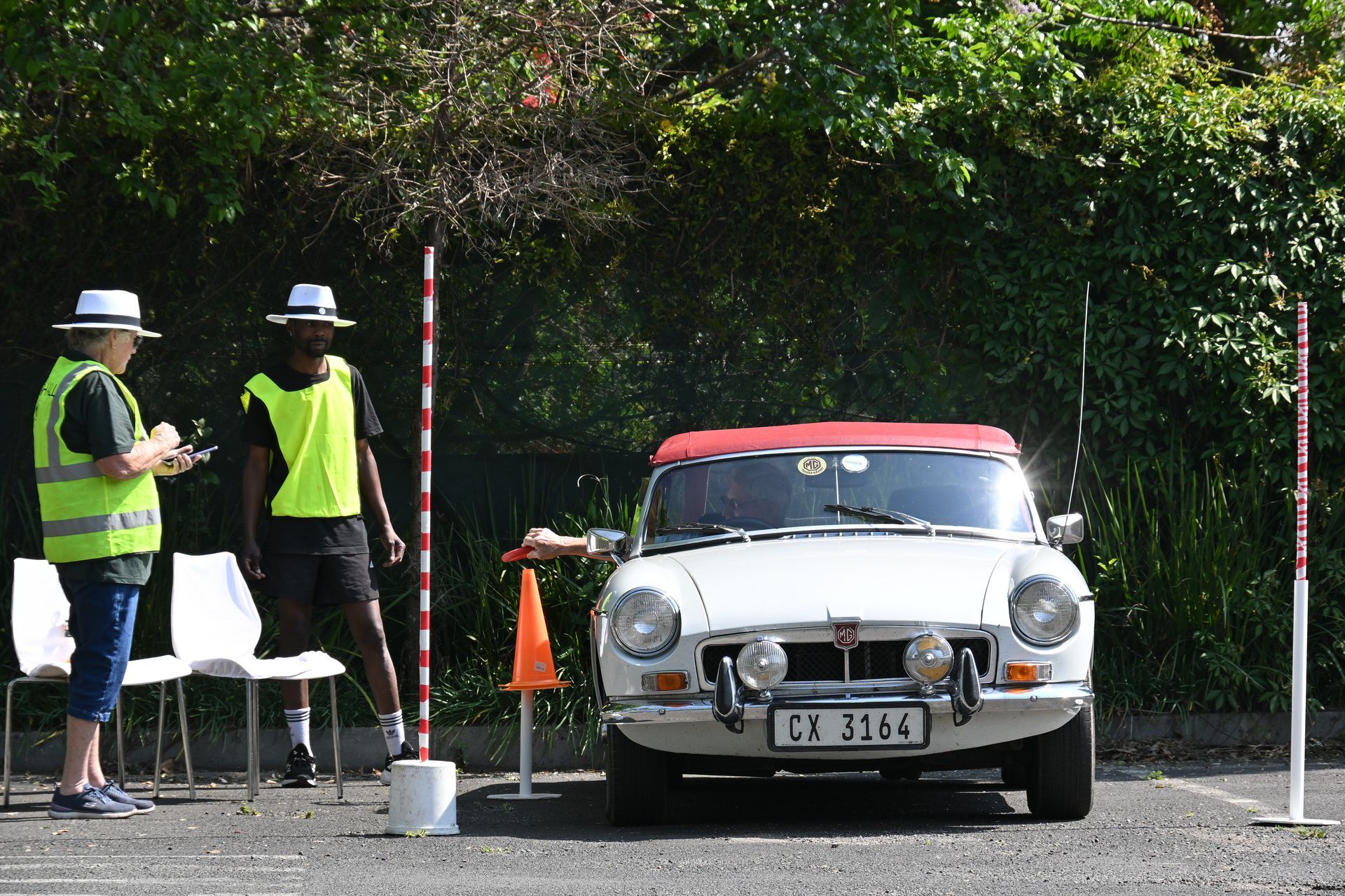 Two workers in safety vests stand beside a classic silver car with a red roof on a roadside course.