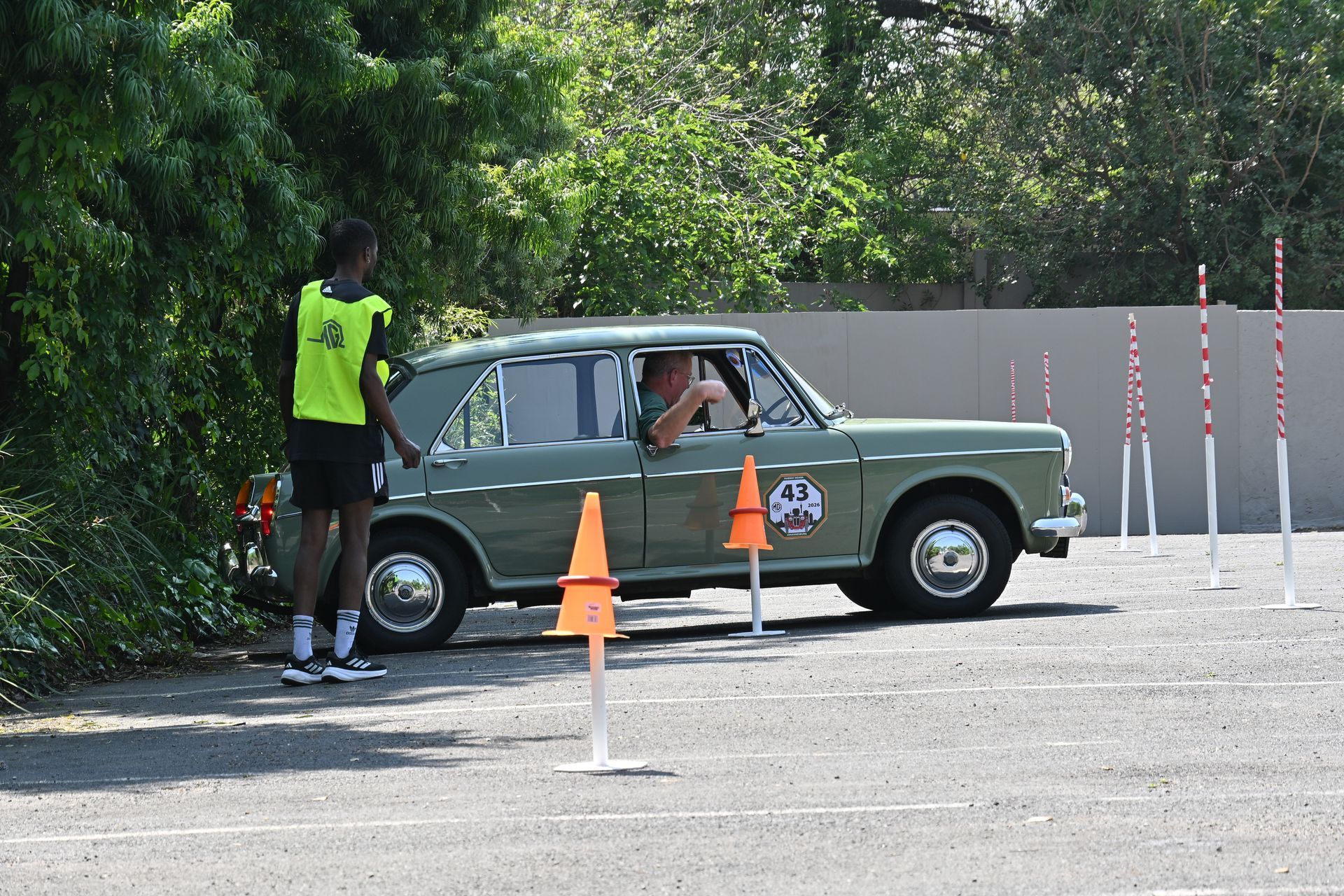 Driver in vintage green car at a roadside checkpoint with a staff member and orange cones.