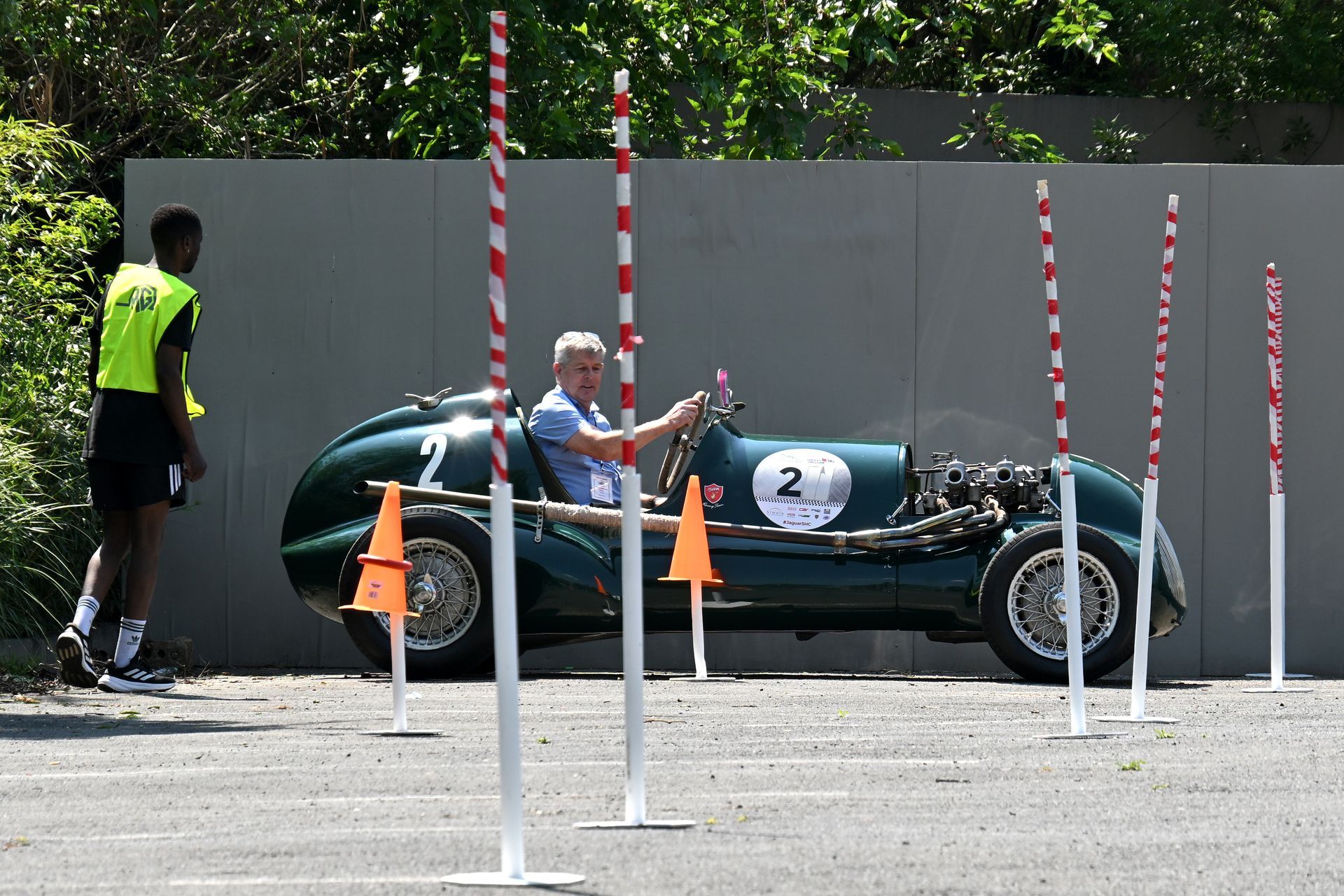 Vintage green race car with driver at a track, beside striped poles and a marshal in a neon vest
