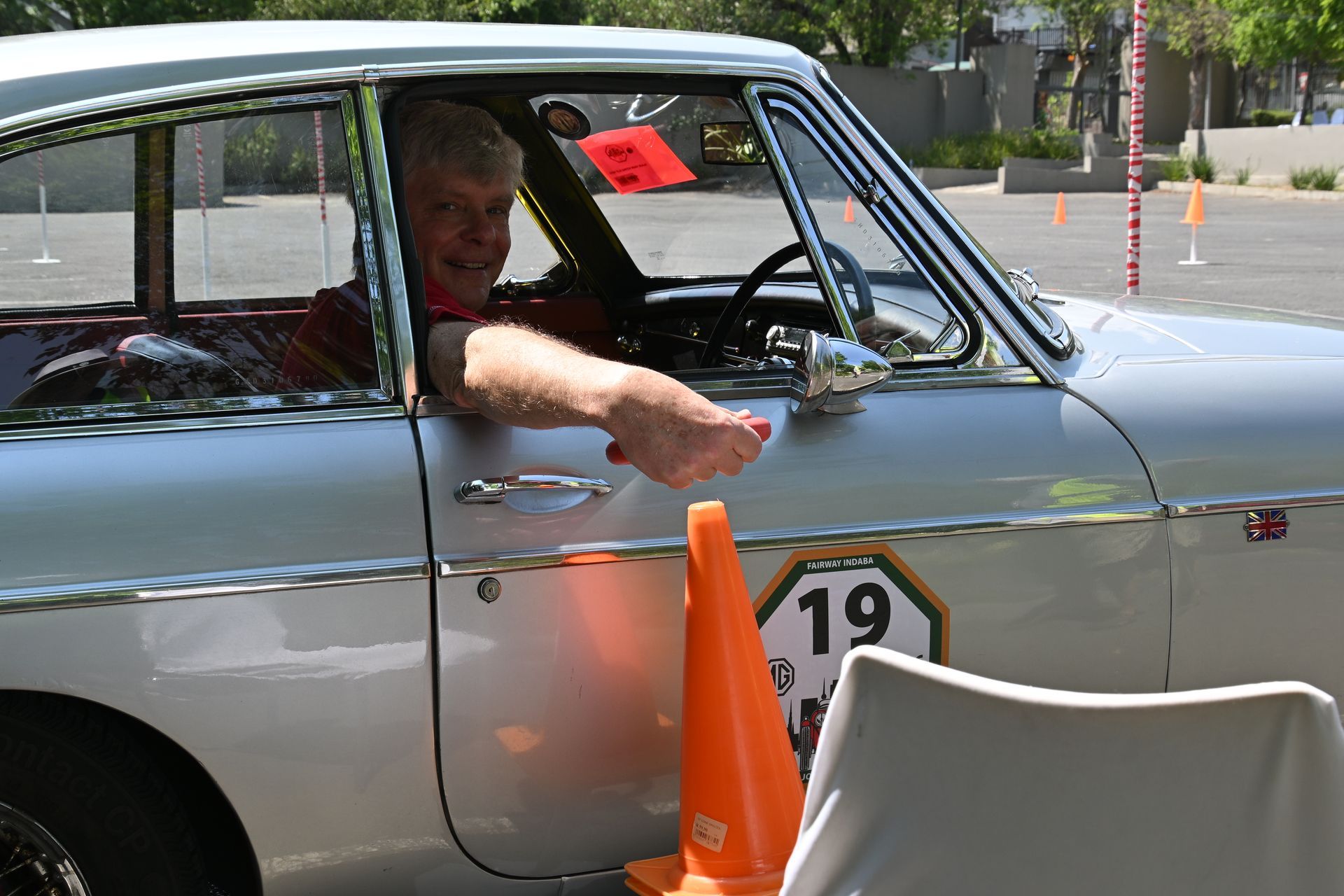 Person in a silver car reaches toward an orange traffic cone near a race course marker 19