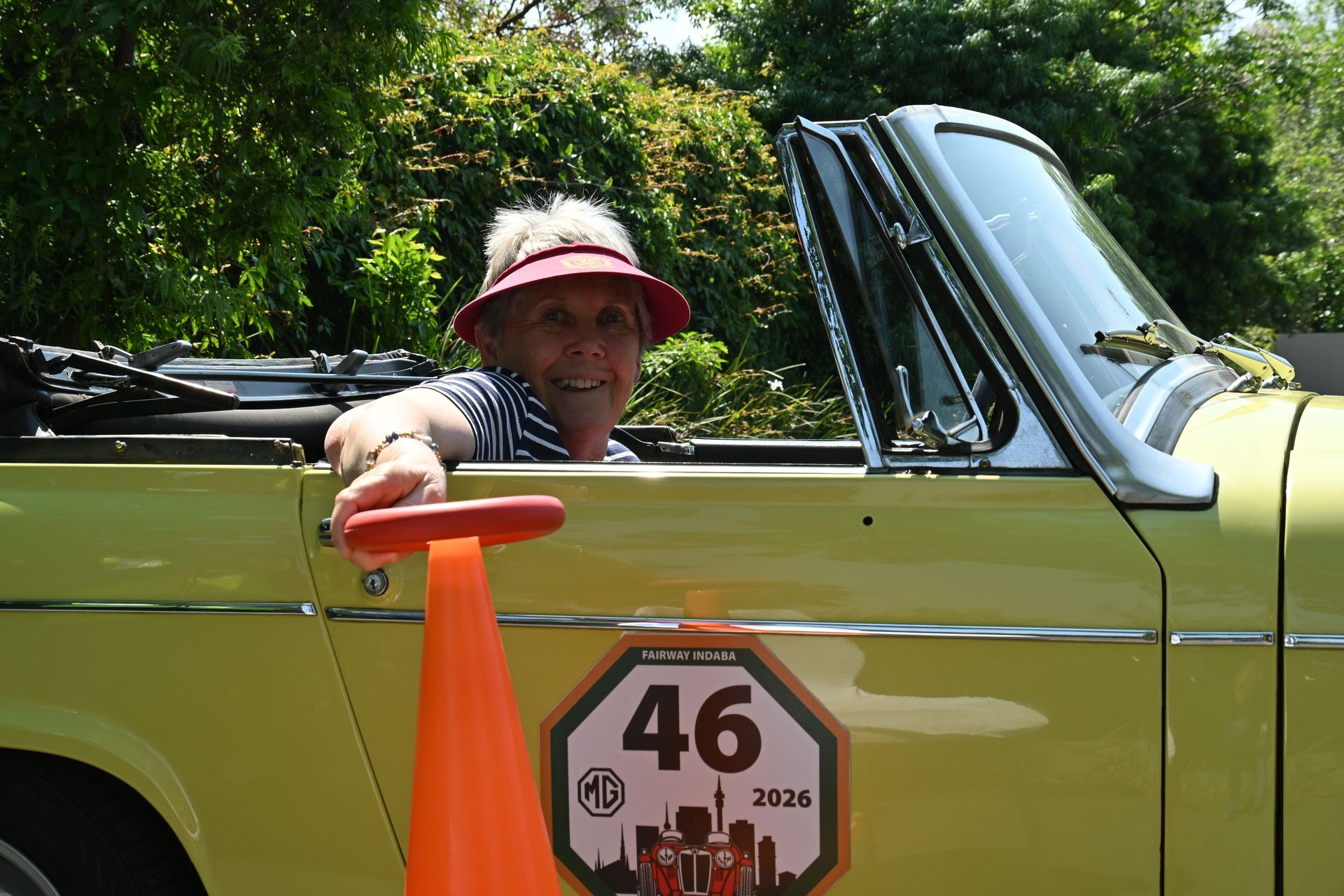 Person smiling in a yellow convertible, wearing a pink hat, with a race number 46 on the door.