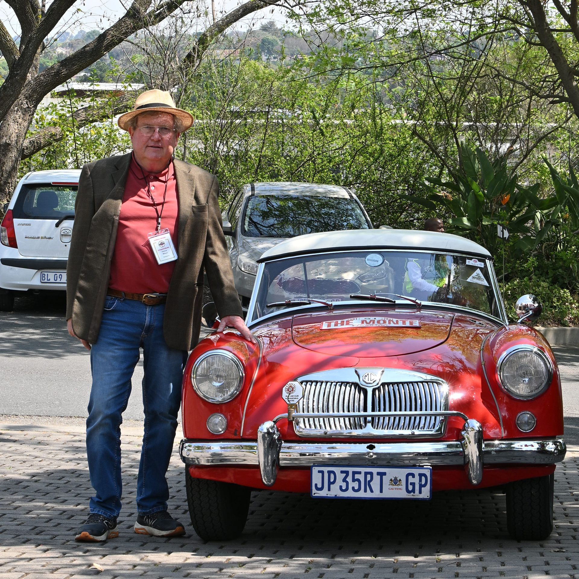 Man standing beside a red vintage car with a white roof on a tree-lined road