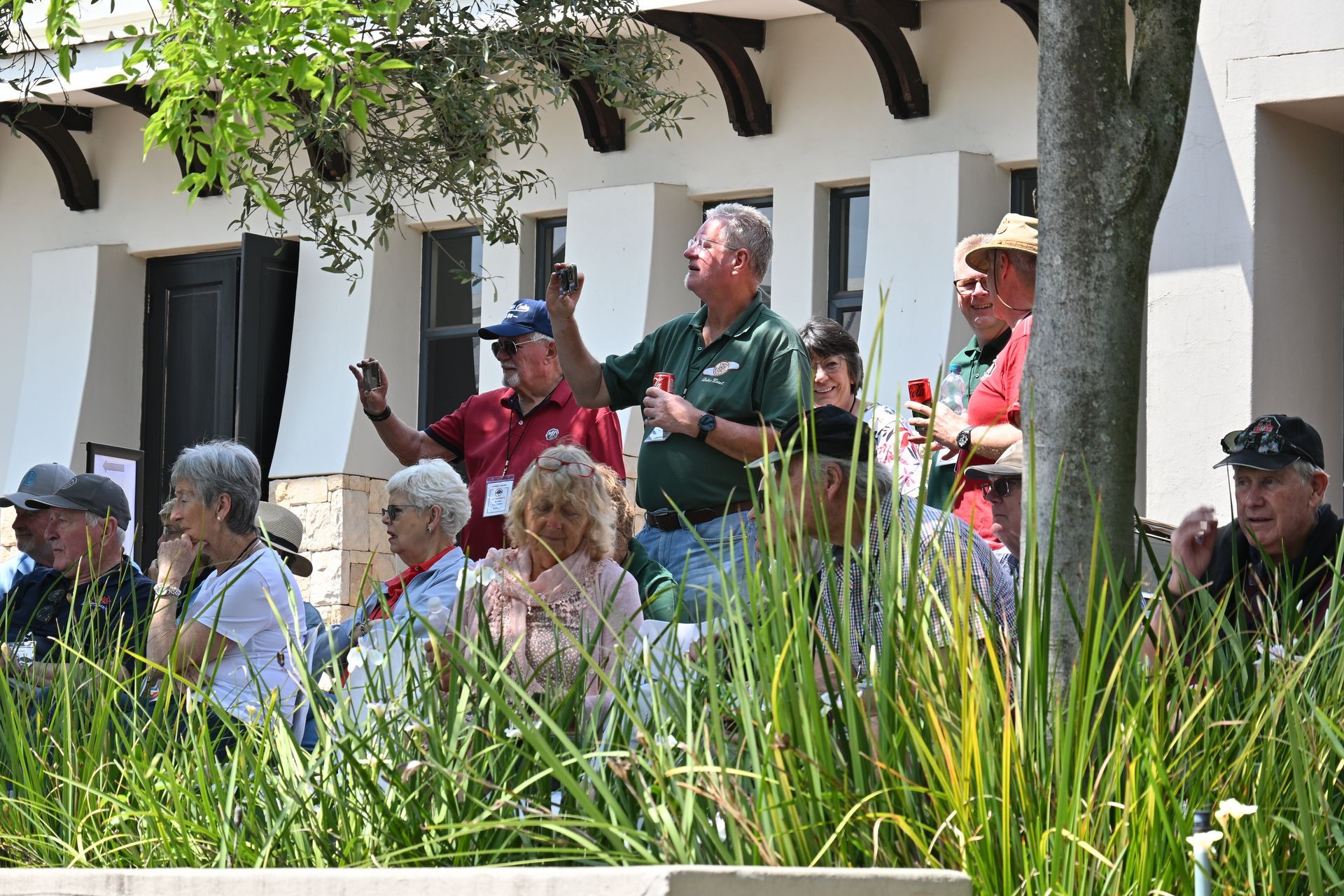 Group of people sitting and standing outdoors by a white building, with tall grass in the foreground