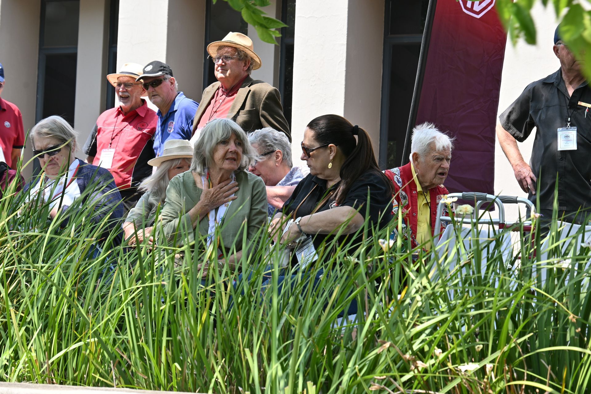 People seated outdoors behind tall grass, watching an event near a building.