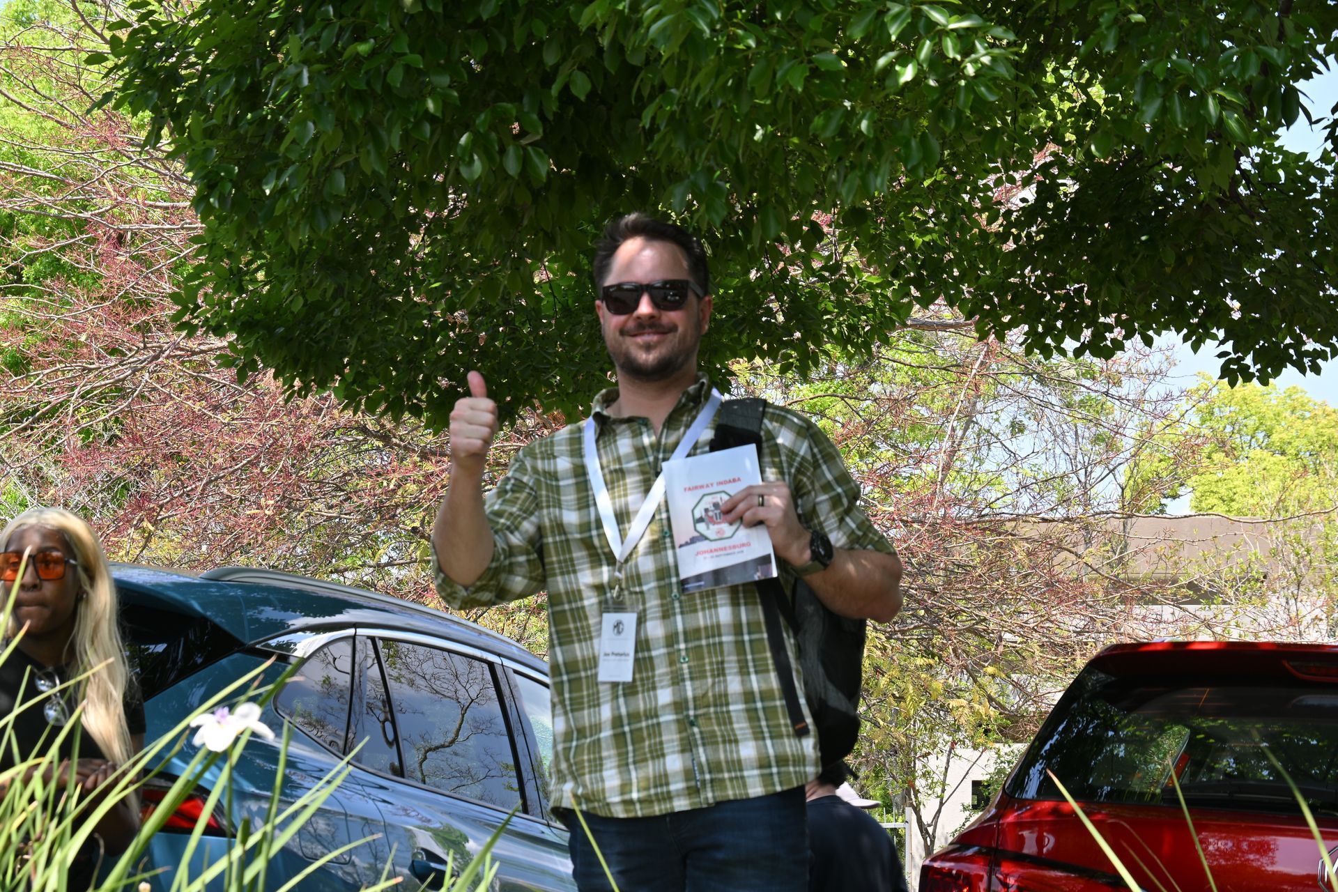 Man standing outdoors under a tree, holding a pamphlet and giving a thumbs-up beside parked cars.
