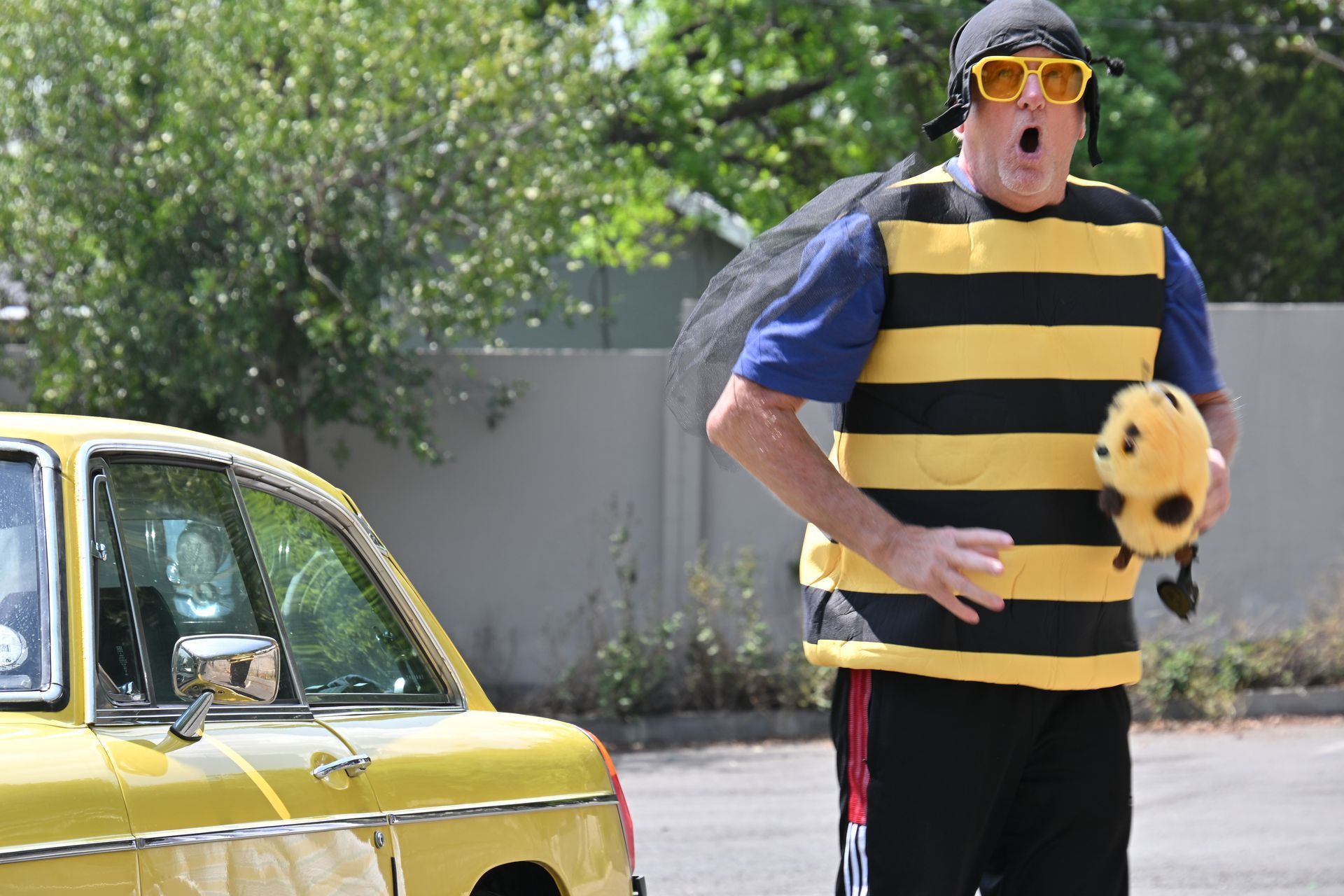 Person in bee costume standing by a yellow car outdoors, holding a plush bee toy.