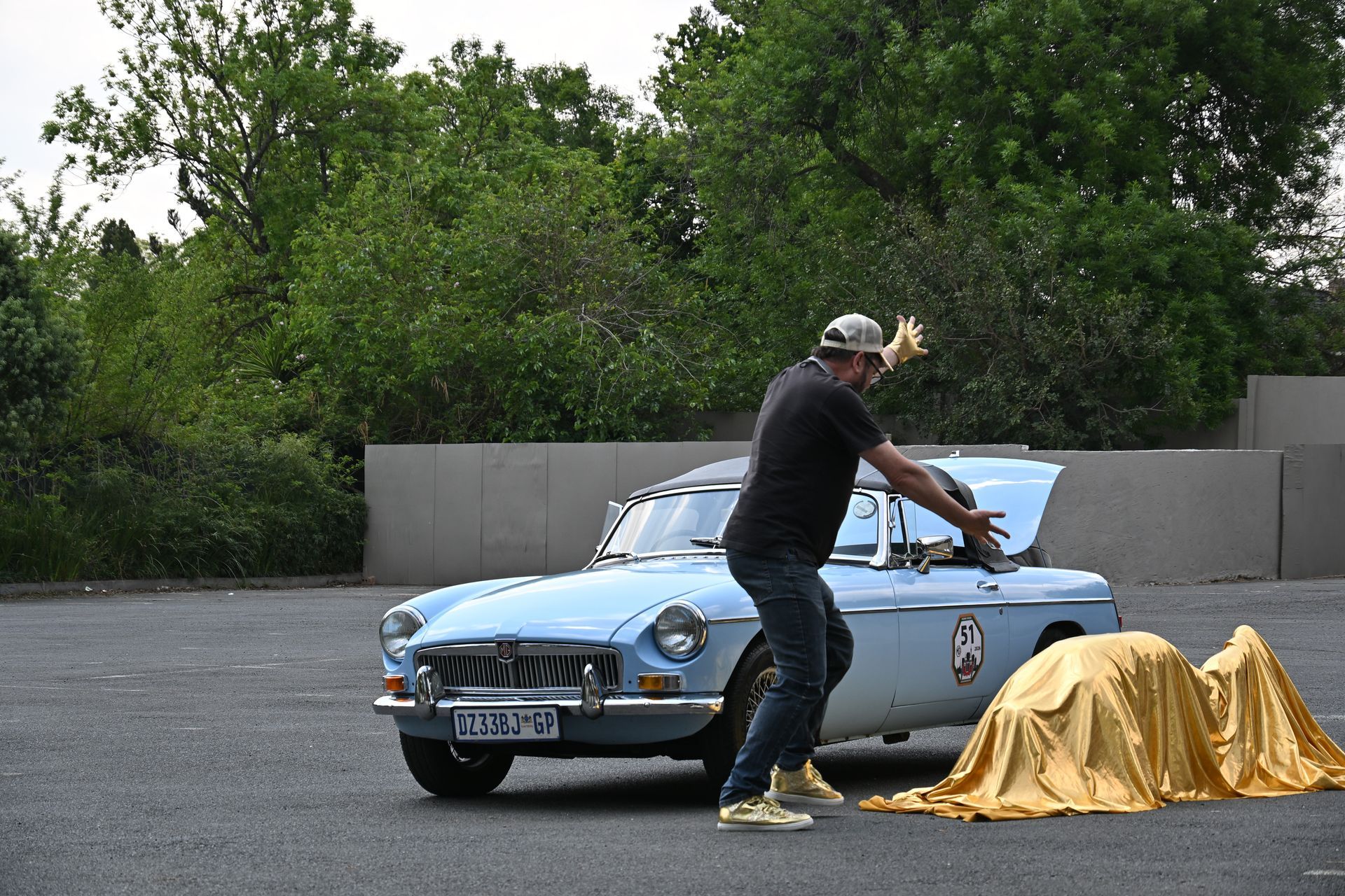 Man pushing a blue vintage car beside a gold-covered object in a parking lot