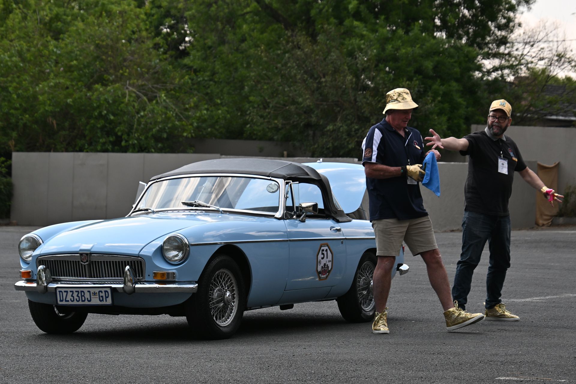 Two men standing beside a light blue vintage convertible on a road, with trees and a wall behind.