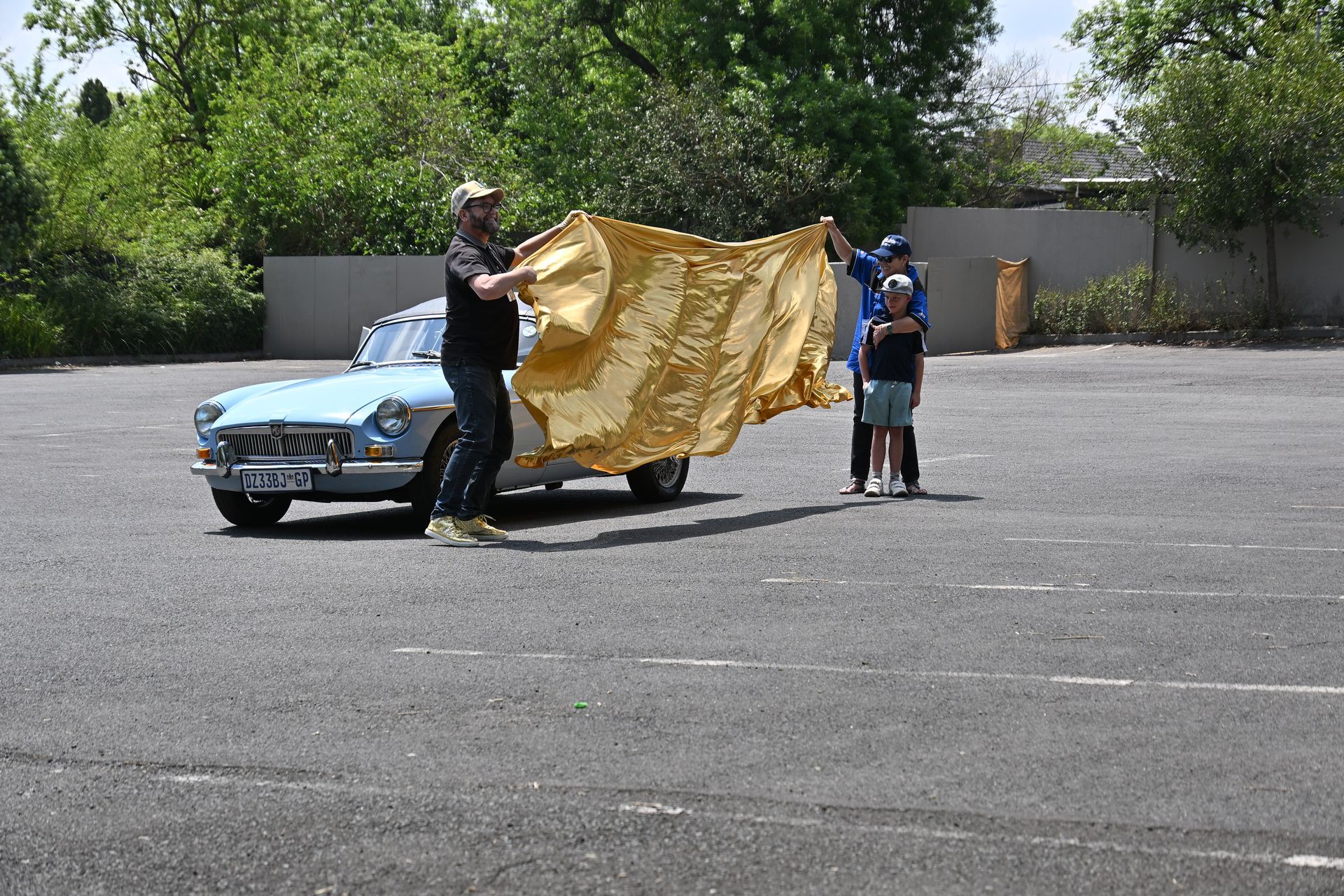 Two people hold up a gold tarp beside a light blue car in an empty parking lot