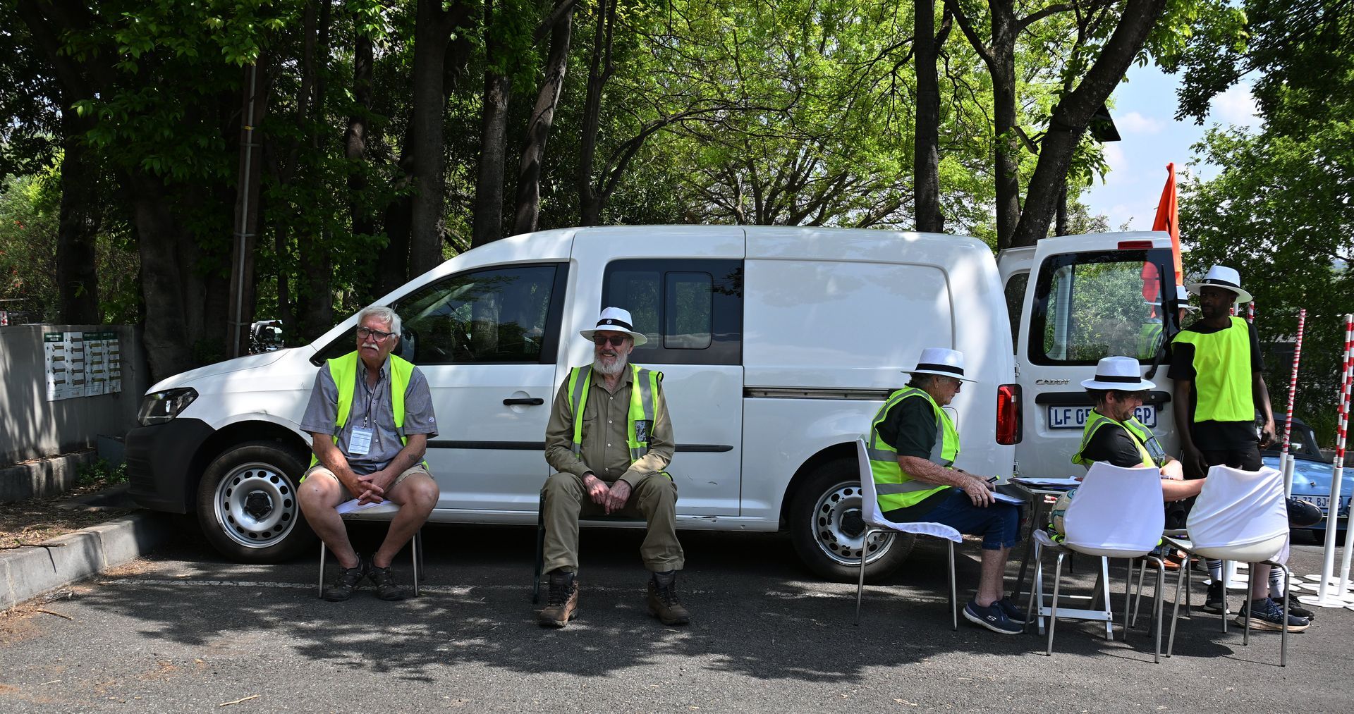 Workers in neon vests beside a white van at a roadside, with chairs and a blue chair under trees