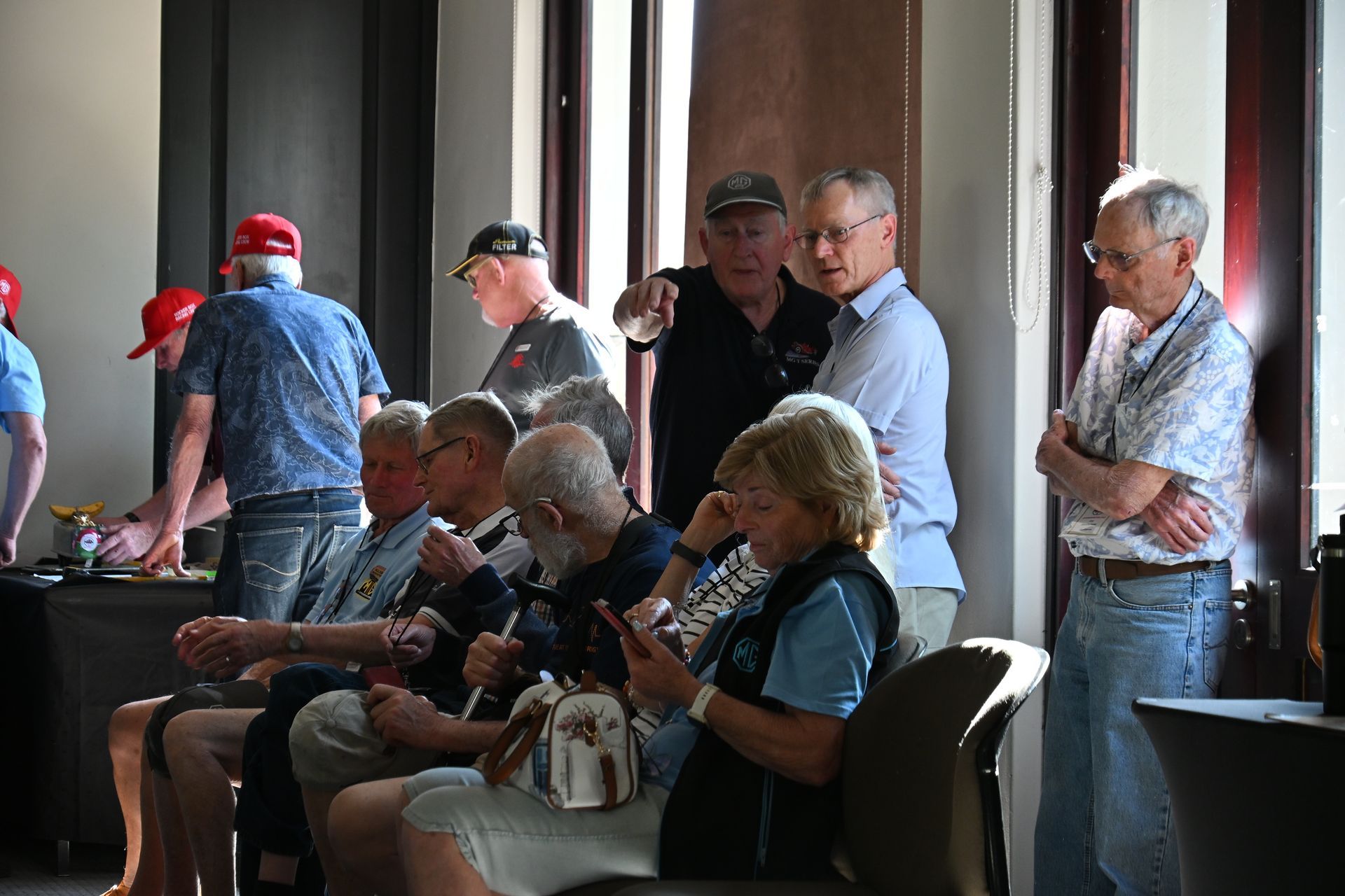 People seated and standing in a dim room, chatting near a table with drinks and a red cap.