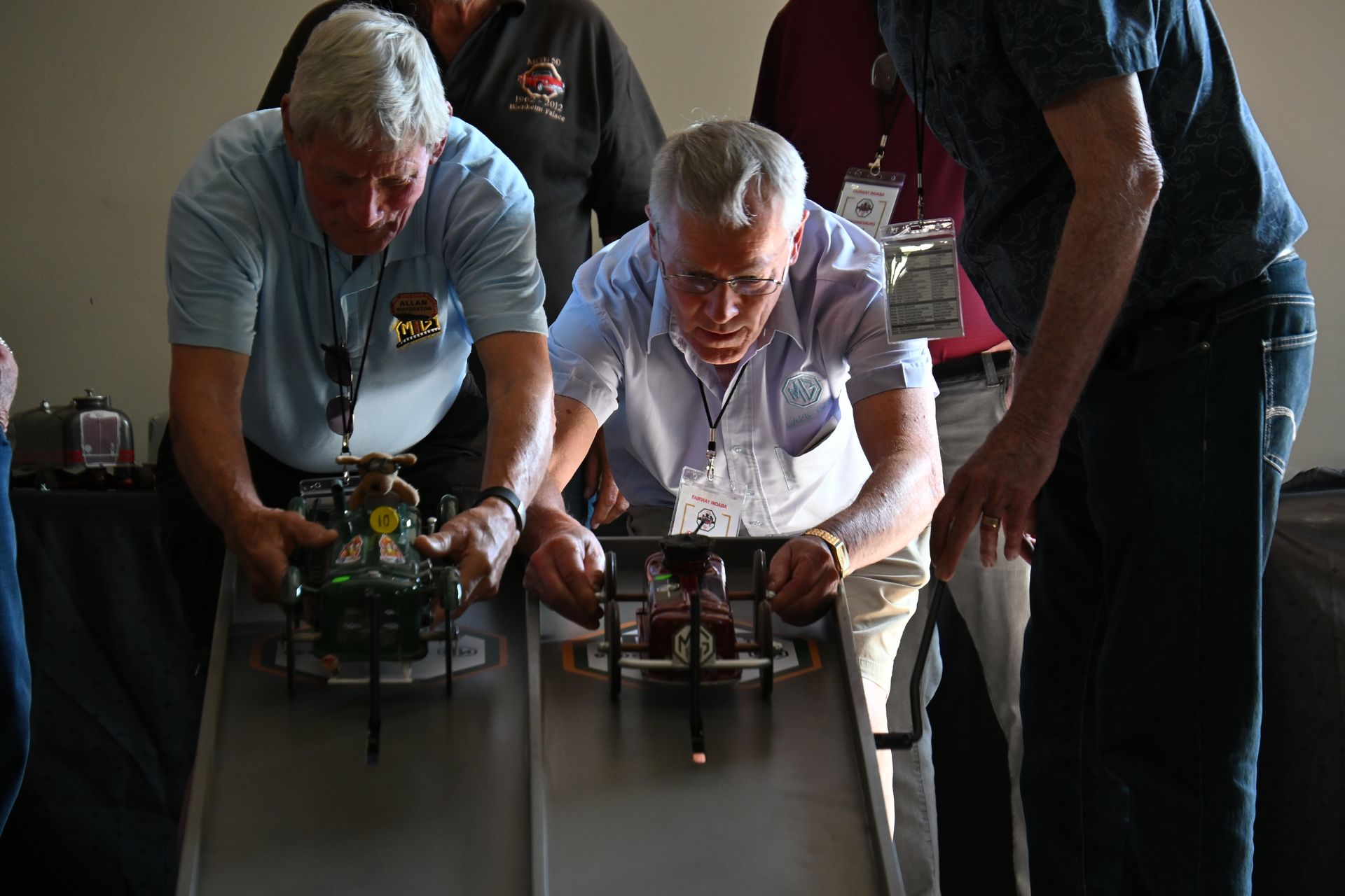 Two men guide small racing cars down adjacent tracks indoors.