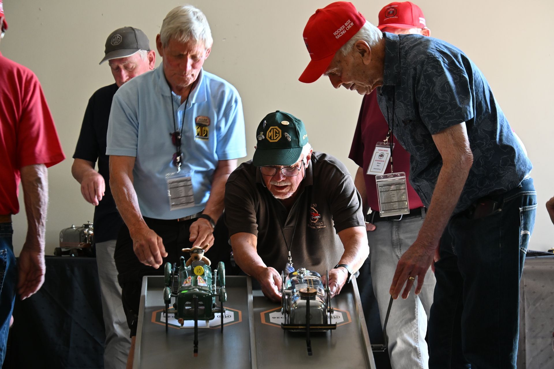 People gathered around small model cars on a table, watching and adjusting them indoors.
