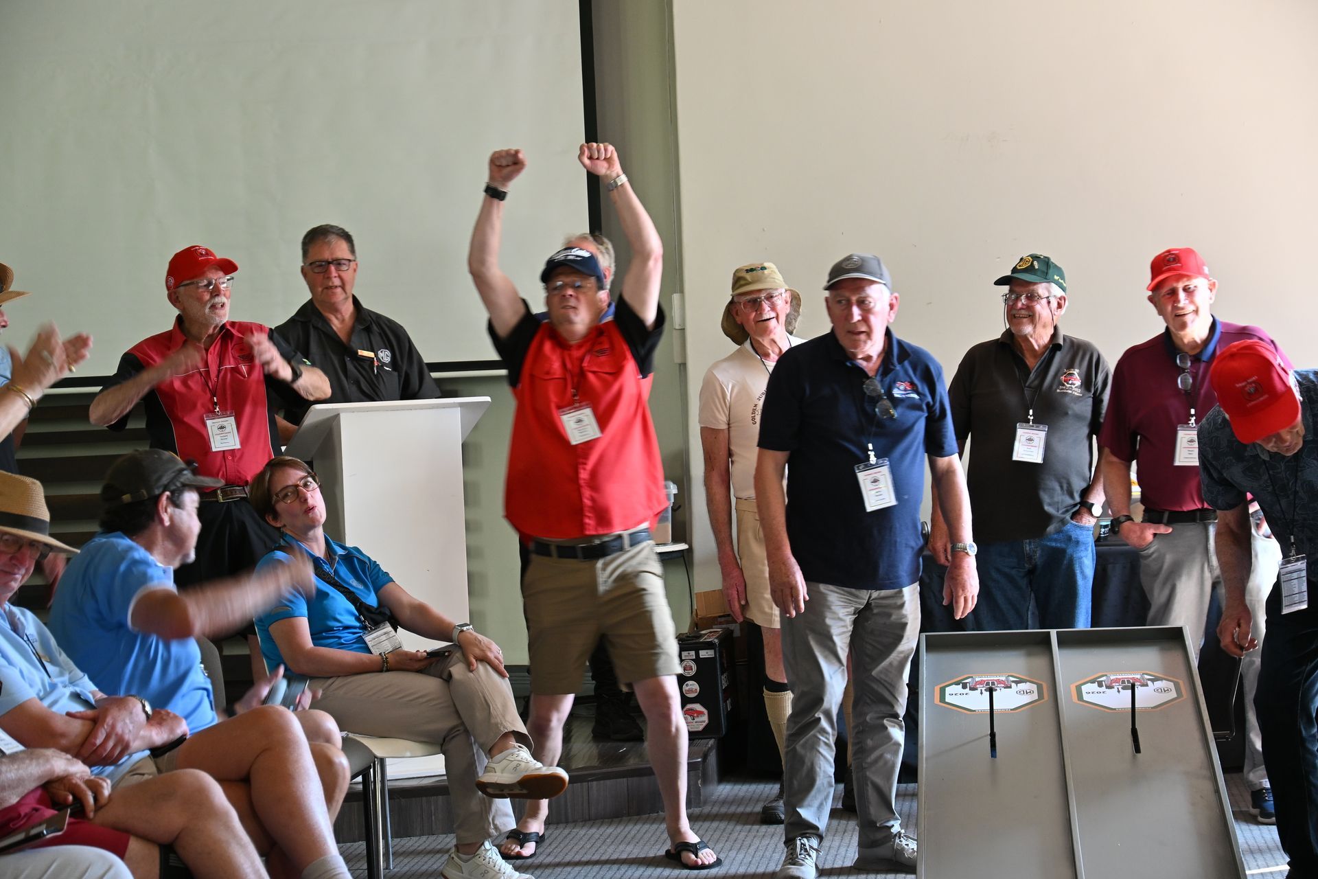 Group of people at an indoor event, some wearing red shirts and hats, cheering with raised arms.