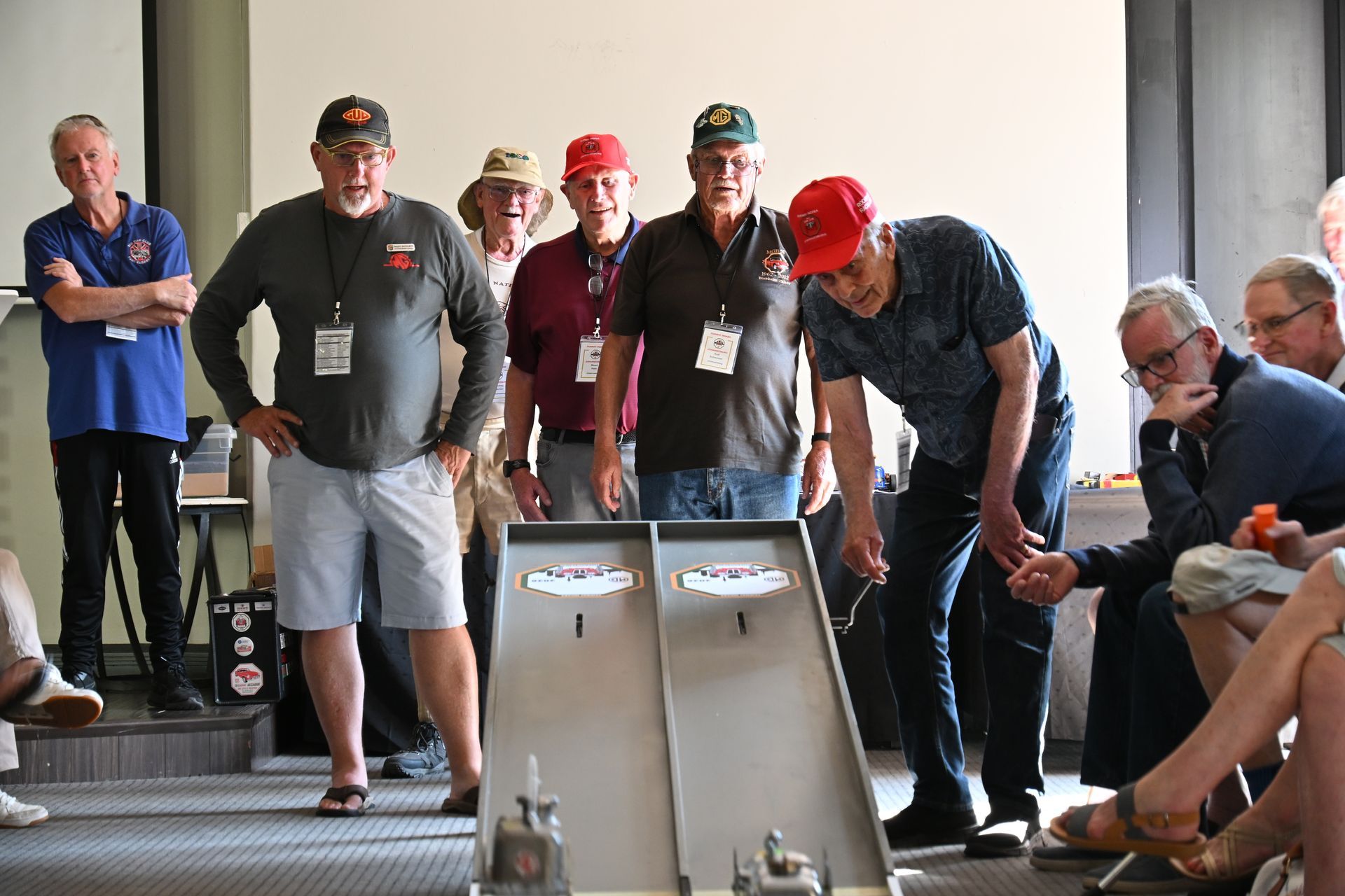 People gathered around a model ramp, watching a sloped track demonstration indoors.