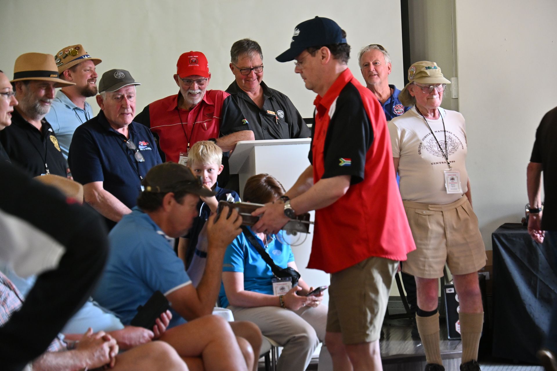Man in red shirt speaking to seated crowd at an indoor event, with photographers and attendees nearby