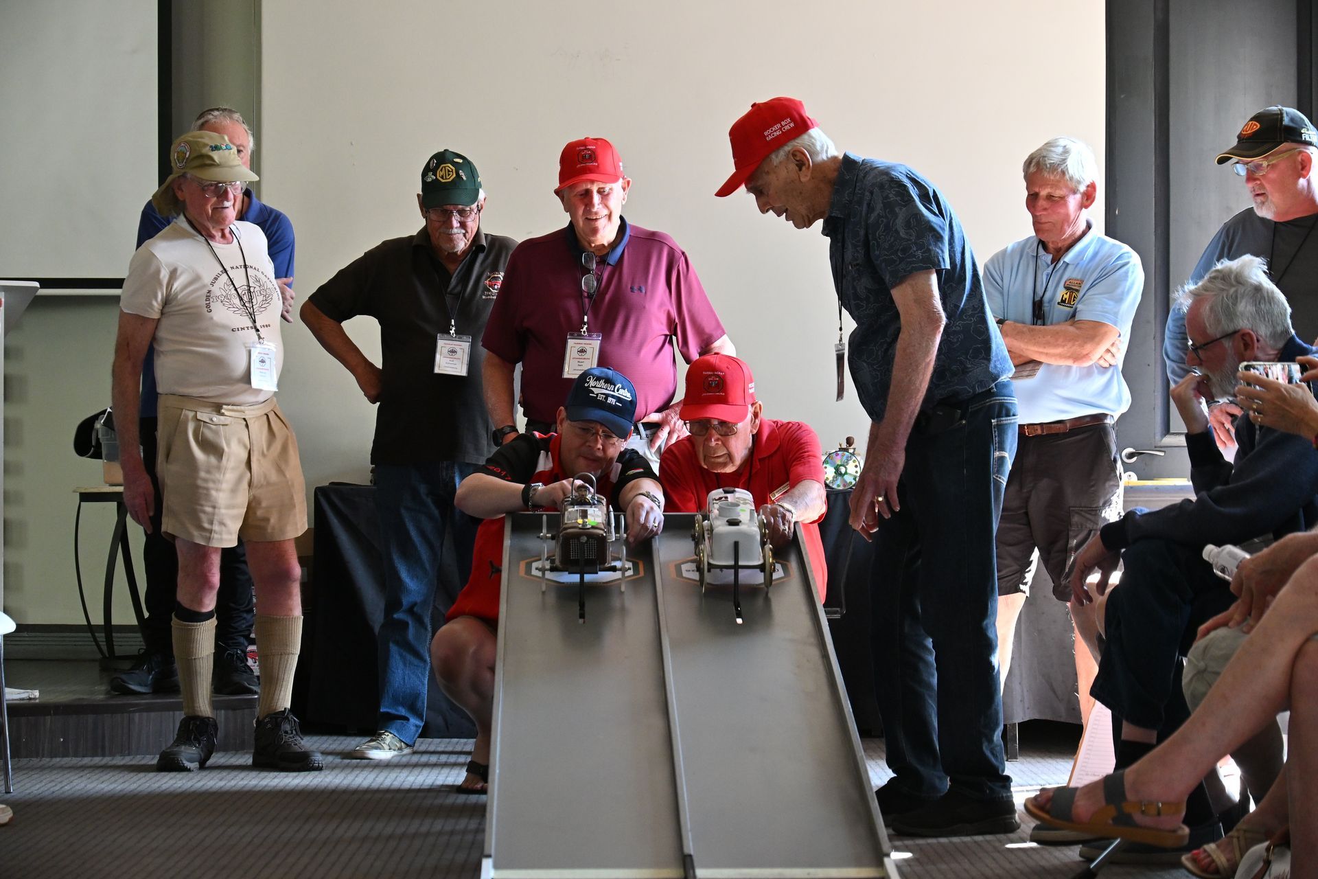 People racing small model cars down a ramp while spectators watch indoors