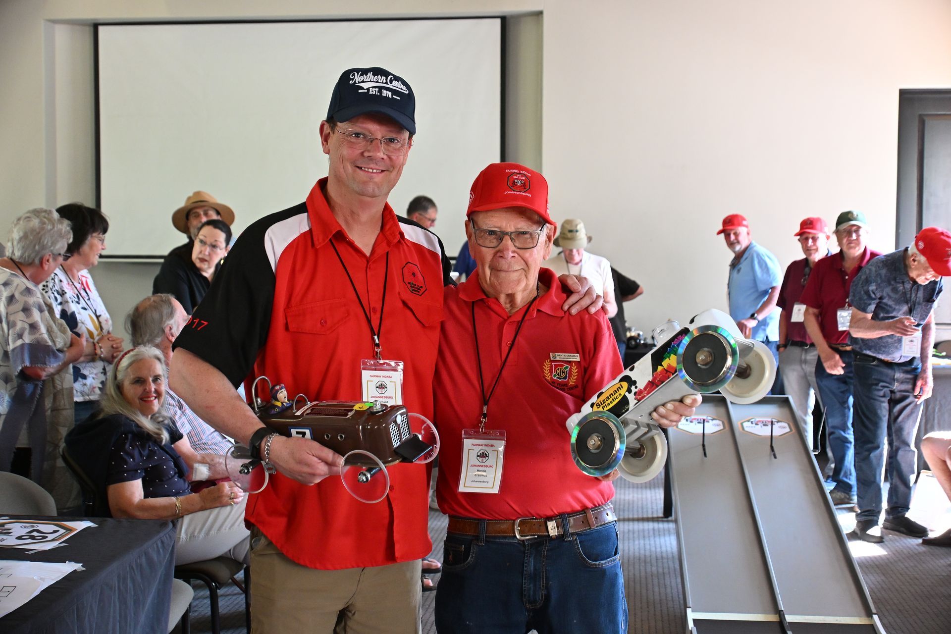 Two people in red shirts and caps holding metal prizes at an indoor event.