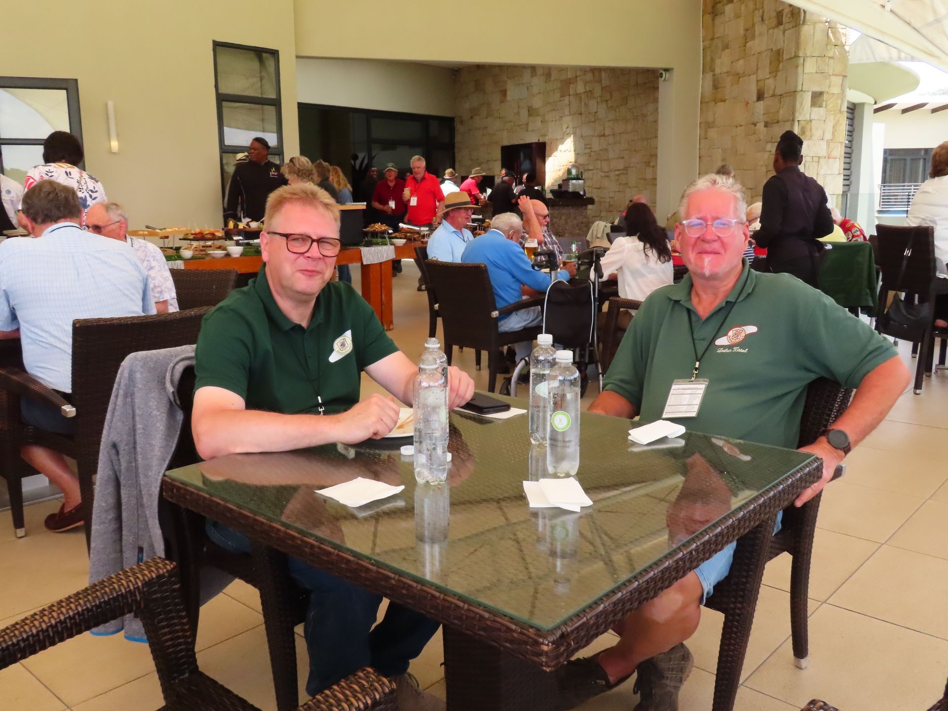 Two people in green shirts sit at a patio table in a busy indoor-outdoor lobby area.