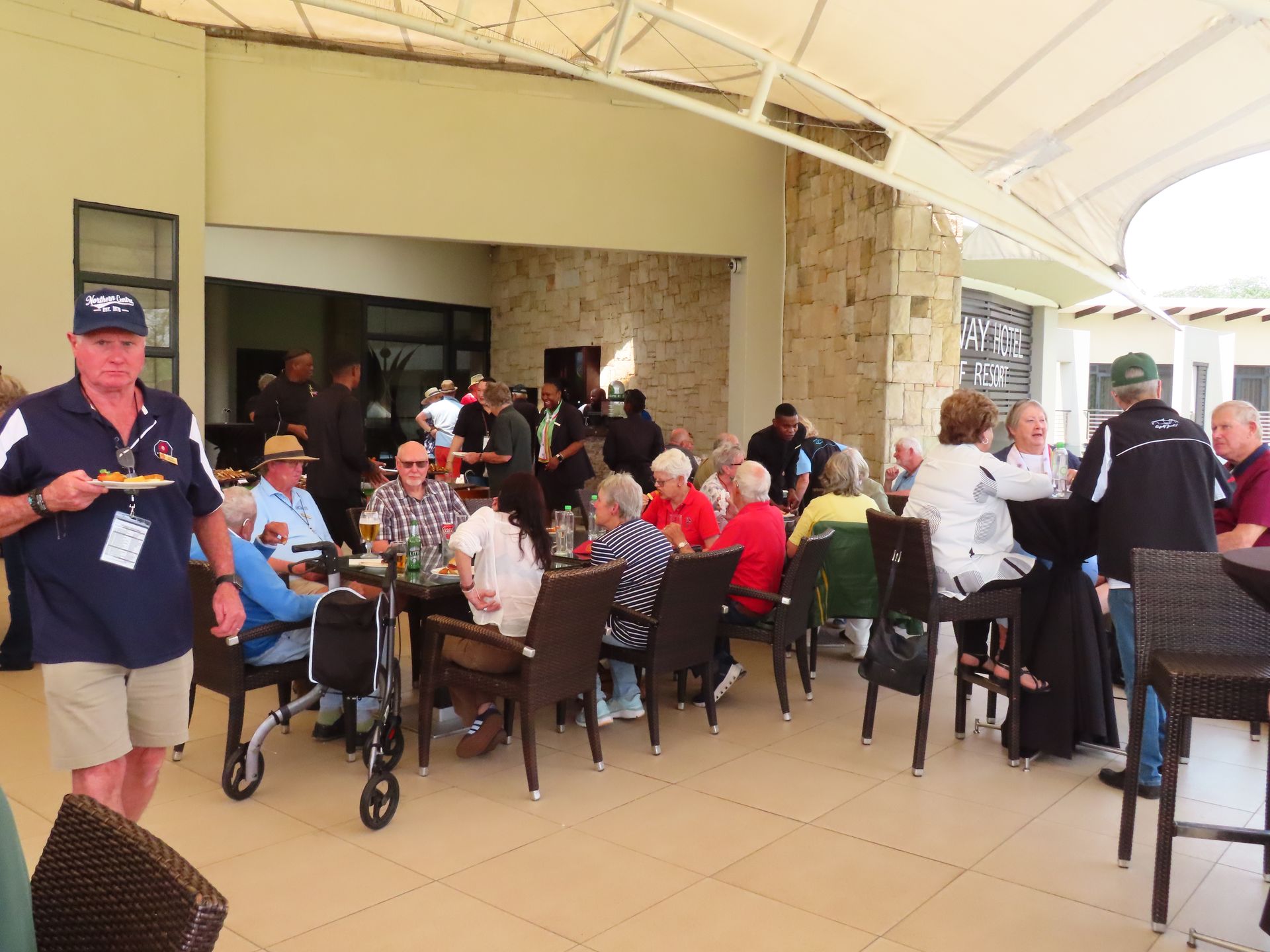 People seated at tables under a covered patio, with a man standing near the left edge.