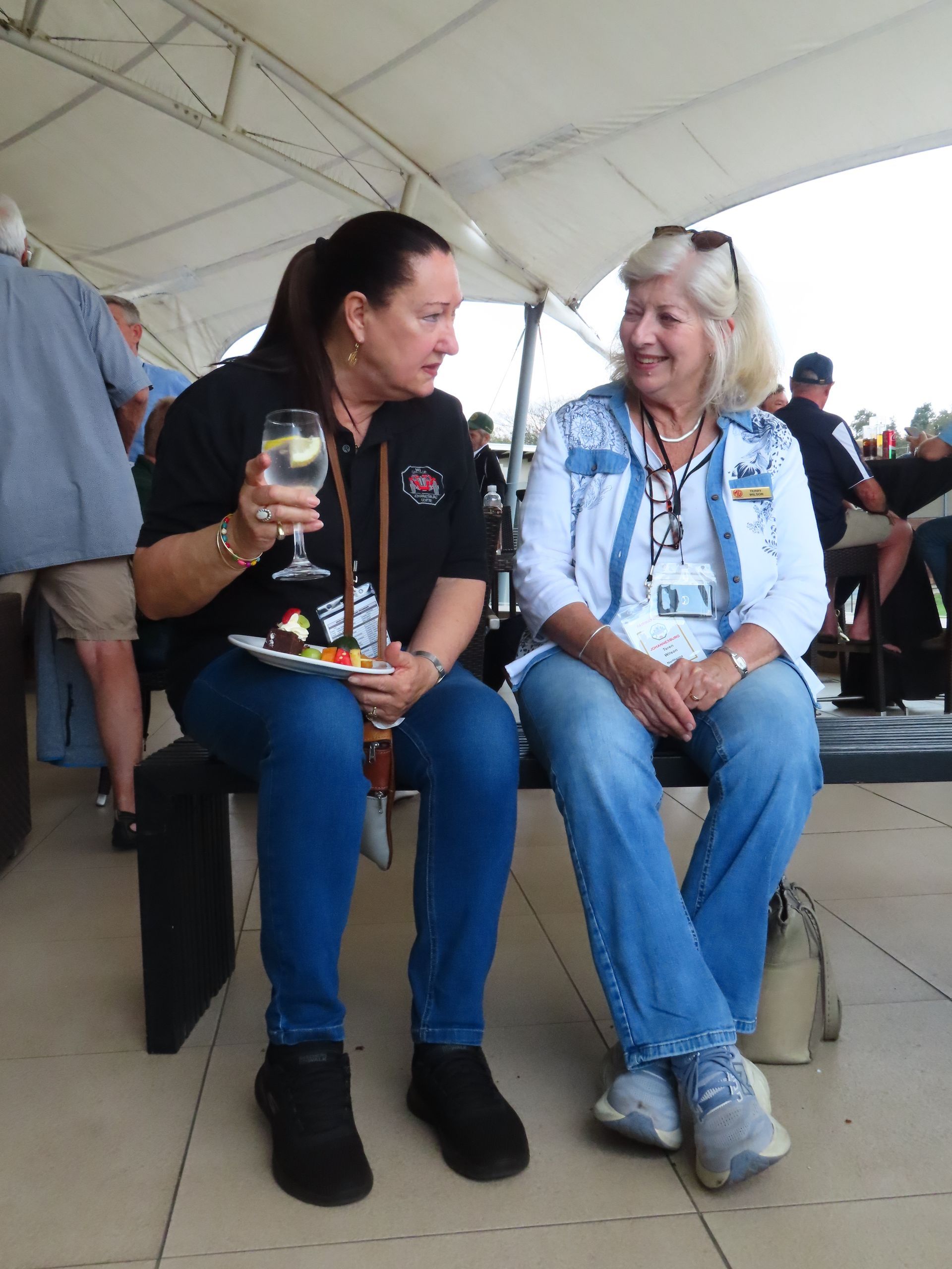 Two people sitting under a white tent, smiling and chatting while holding drinks at an outdoor event