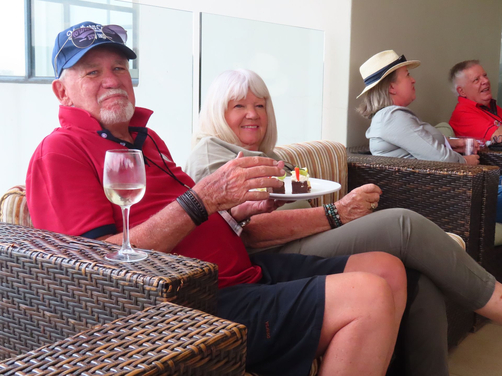 Two people in red shirts sit on wicker chairs on a patio, holding a wine glass and a dessert plate.