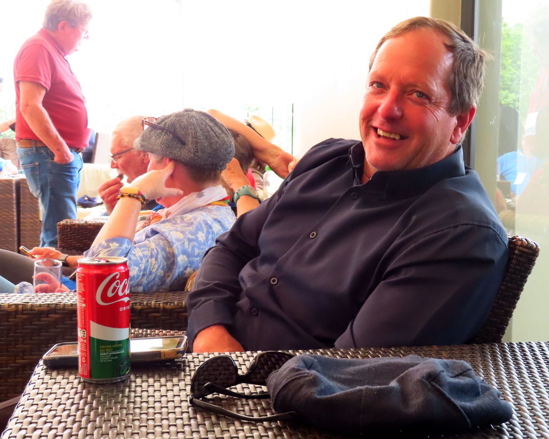 Smiling man in black shirt seated at a table with a Coca-Cola can in a cafe