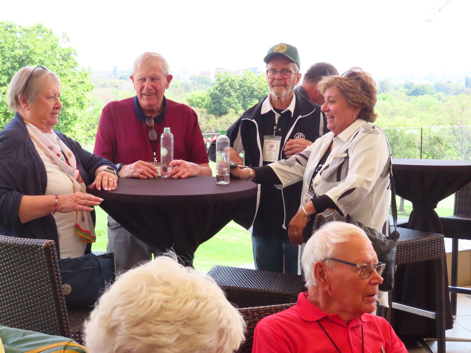 Five people chatting at a round table on a covered patio with drinks, overlooking a green outdoor view