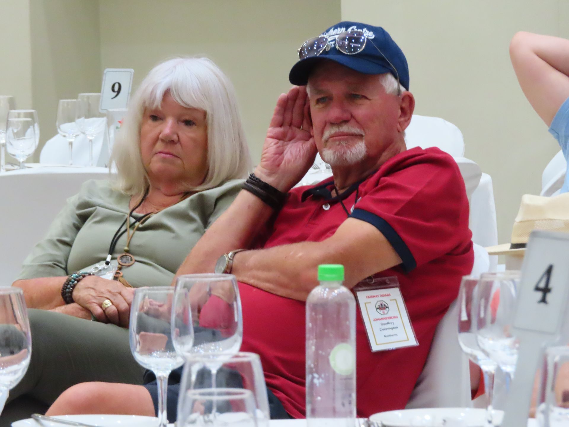 Two people seated at a table at an indoor event, one in a red shirt and cap, the other in a light green top.
