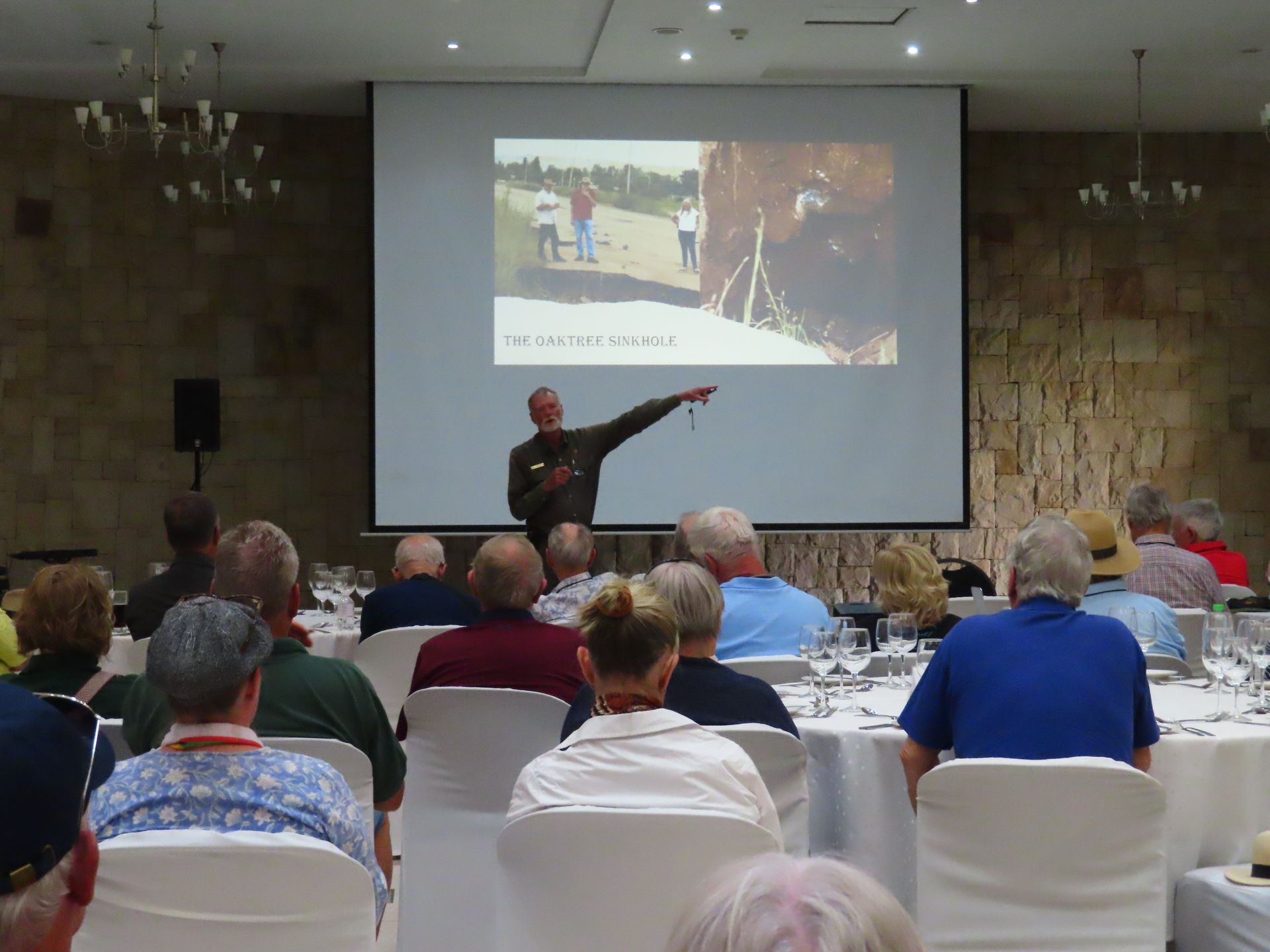 Presenter points at projected slides during a lecture to an audience seated in a conference room