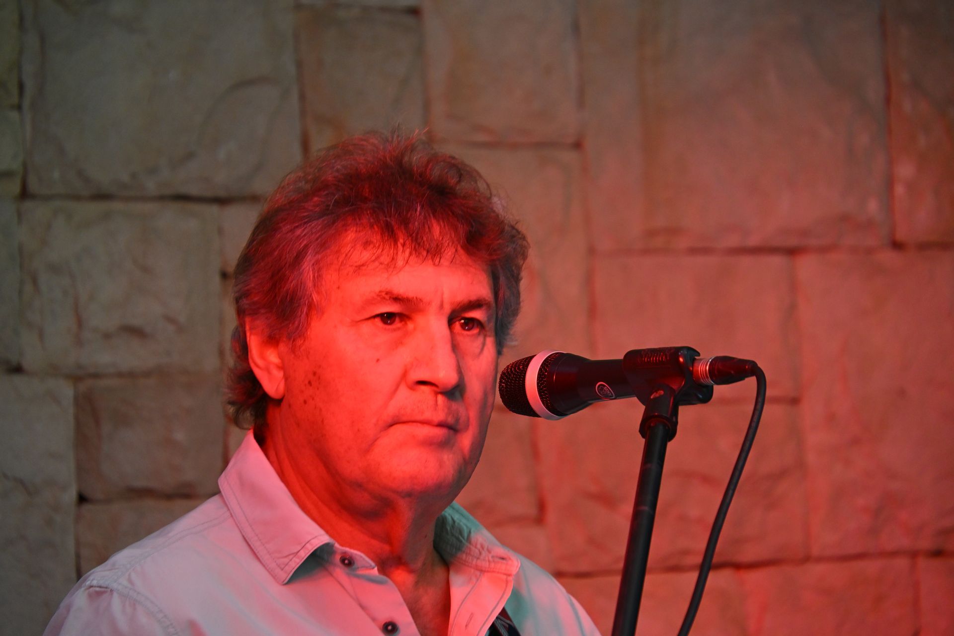 Man singing into a microphone under red stage lighting, with a stone wall backdrop