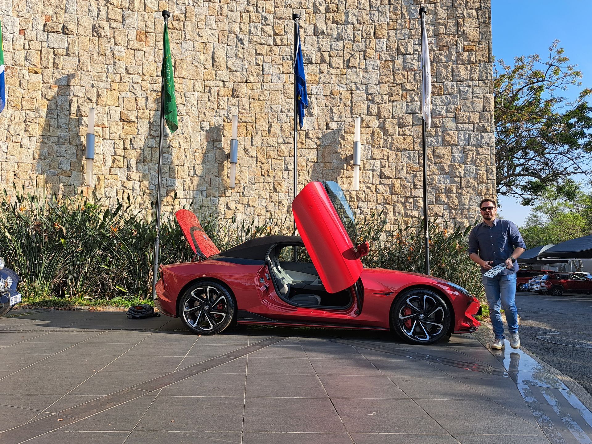 Red sports car with scissor doors open parked by a stone wall; man stands beside it.