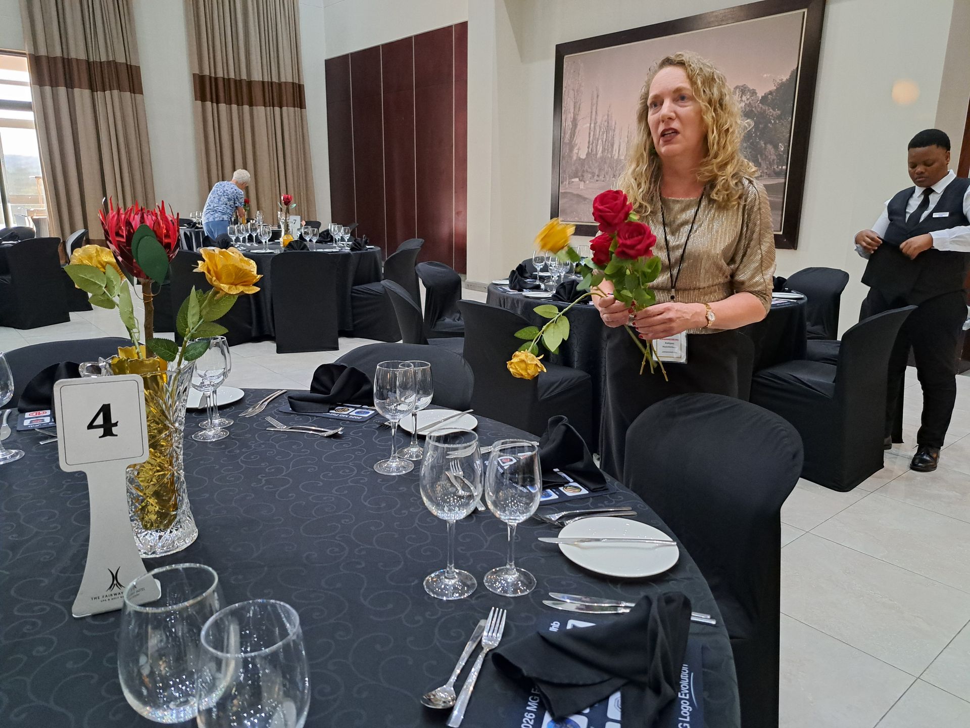 Formal dining table with floral centerpiece and table number 4; woman stands nearby holding red flowers.