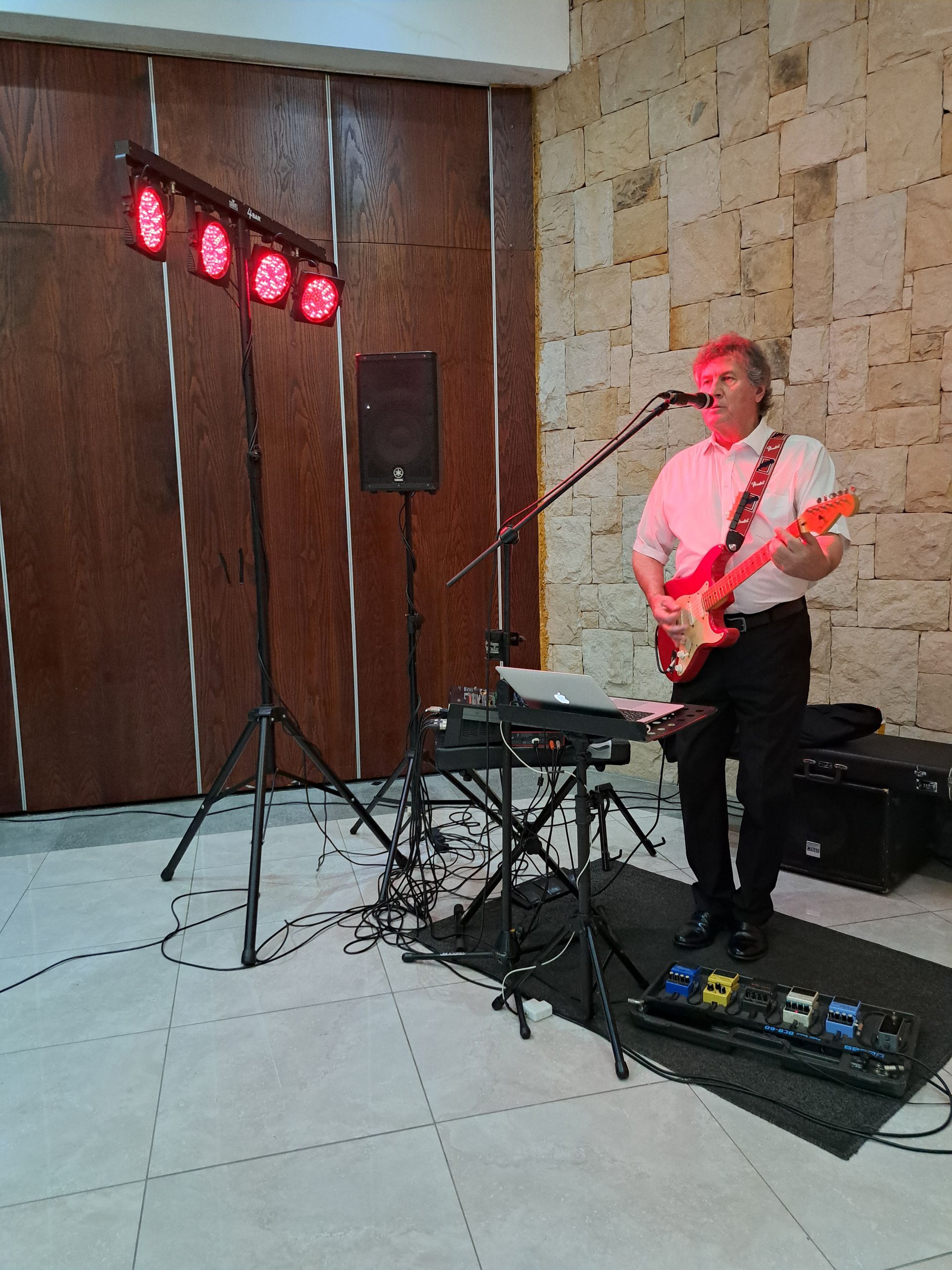 Musician playing electric guitar beside a keyboard and red stage lights in a hall
