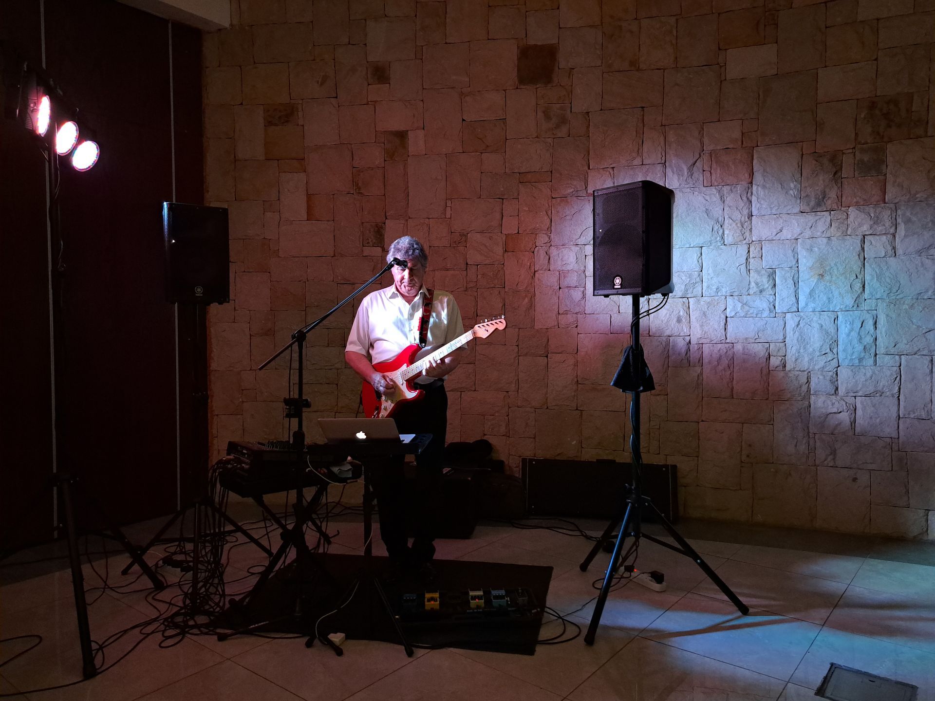 Musician playing electric guitar on a small stage with speakers and microphones against a stone wall.