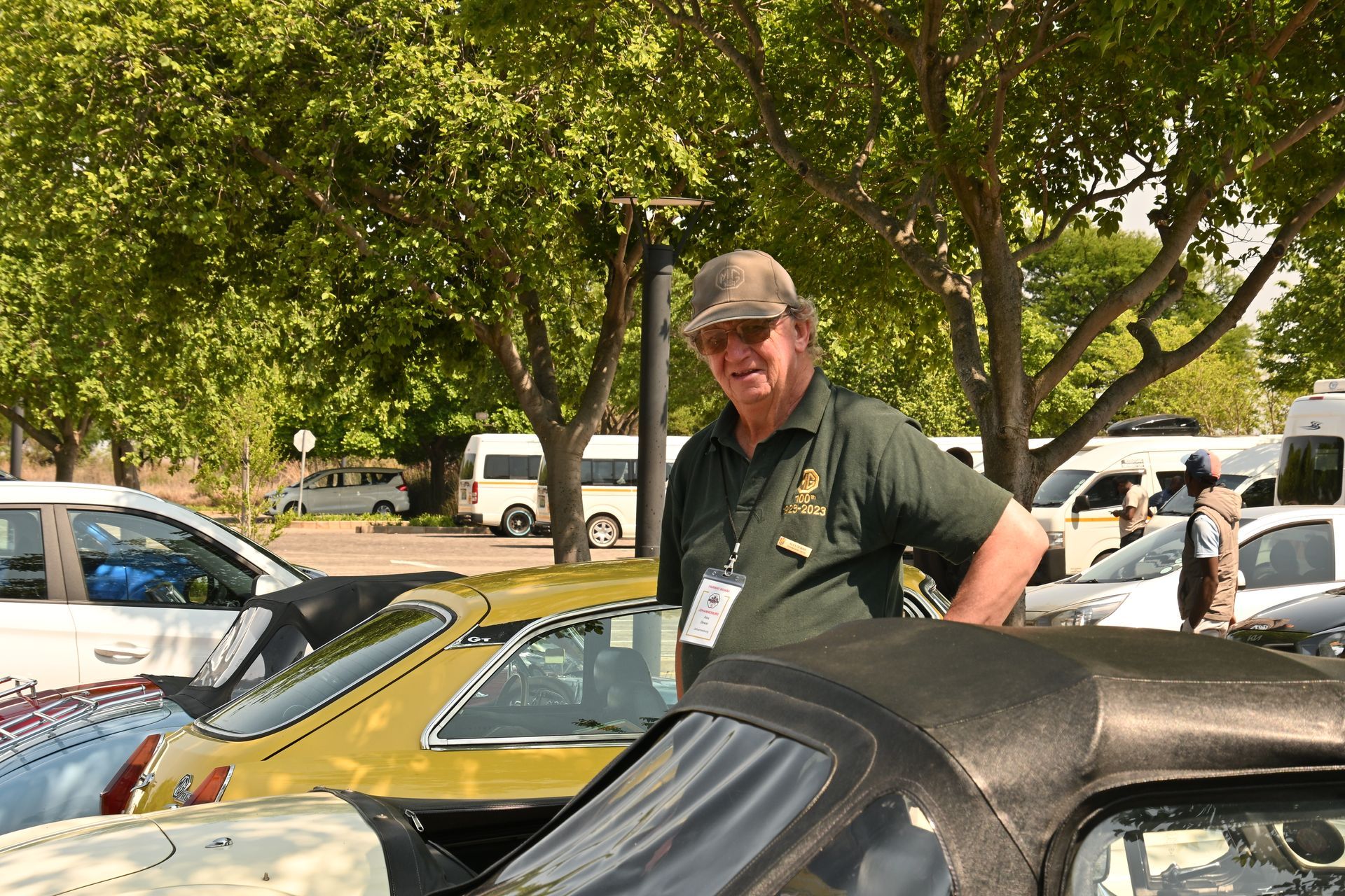 Man smiling beside classic cars at an outdoor car show under trees