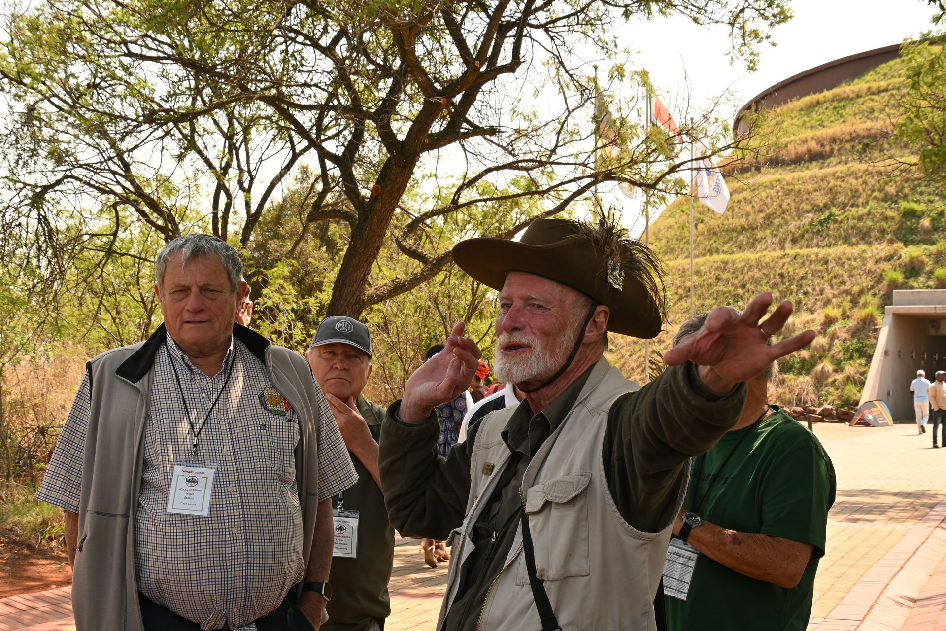 Tour guide in a cowboy hat speaking outdoors with two people near a tree and hillside path